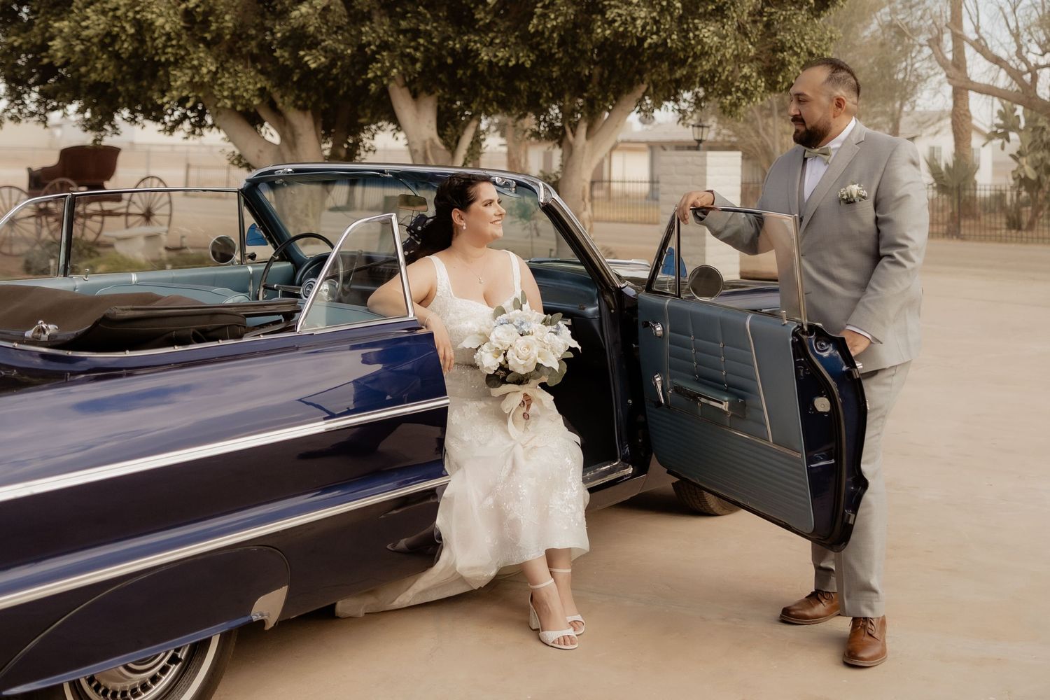 Bride in lace wedding dress steps out of a classic navy blue vintage car as groom in gray suit holds the door.