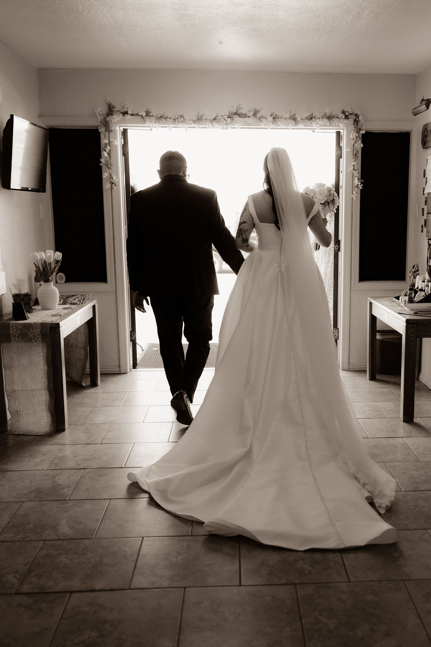 Black and white photo of bride and groom walking hand in hand toward bright doorway after wedding.