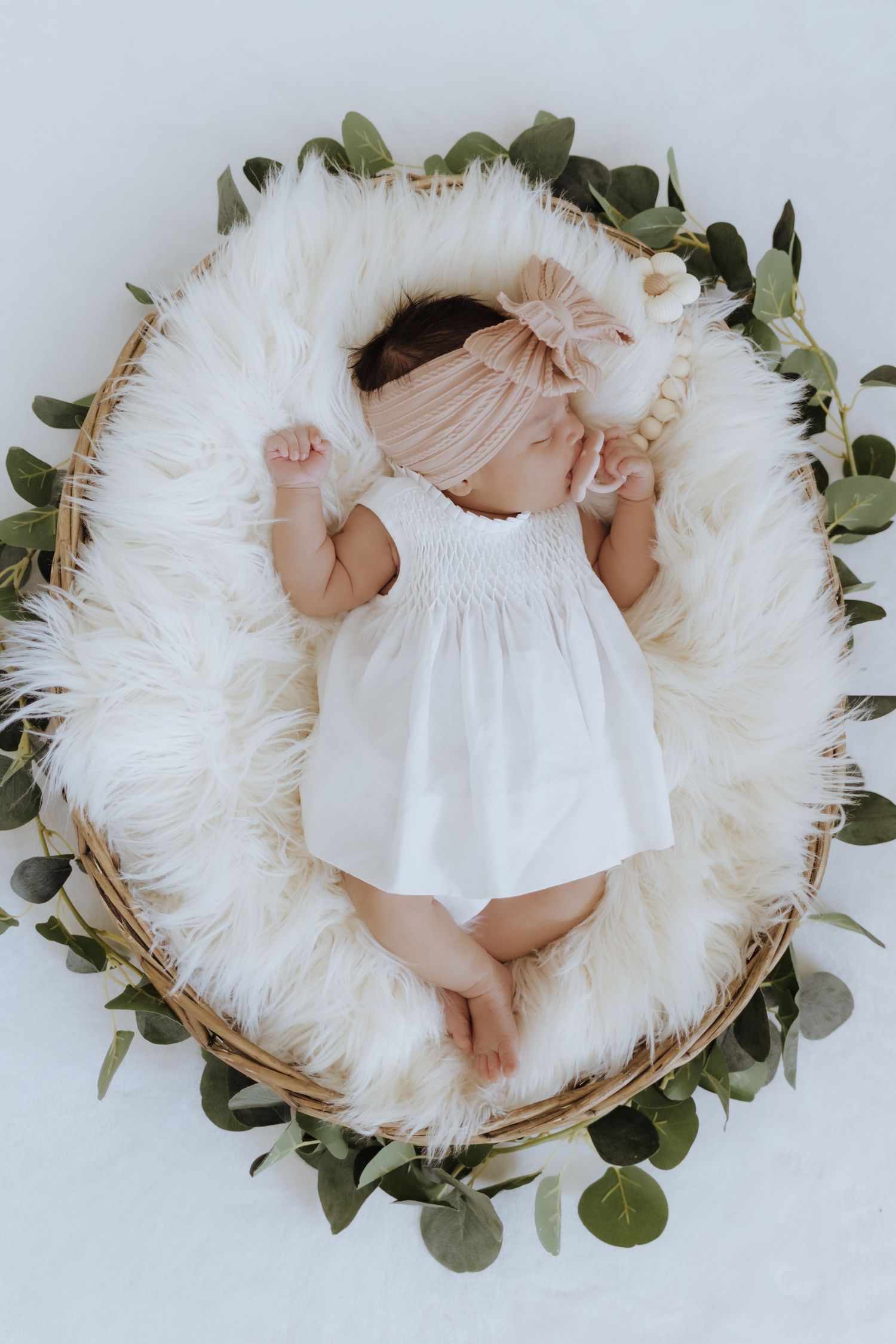 Newborn baby sleeping in a wicker basket with white fur lining, wearing a white dress and pink bow headband.