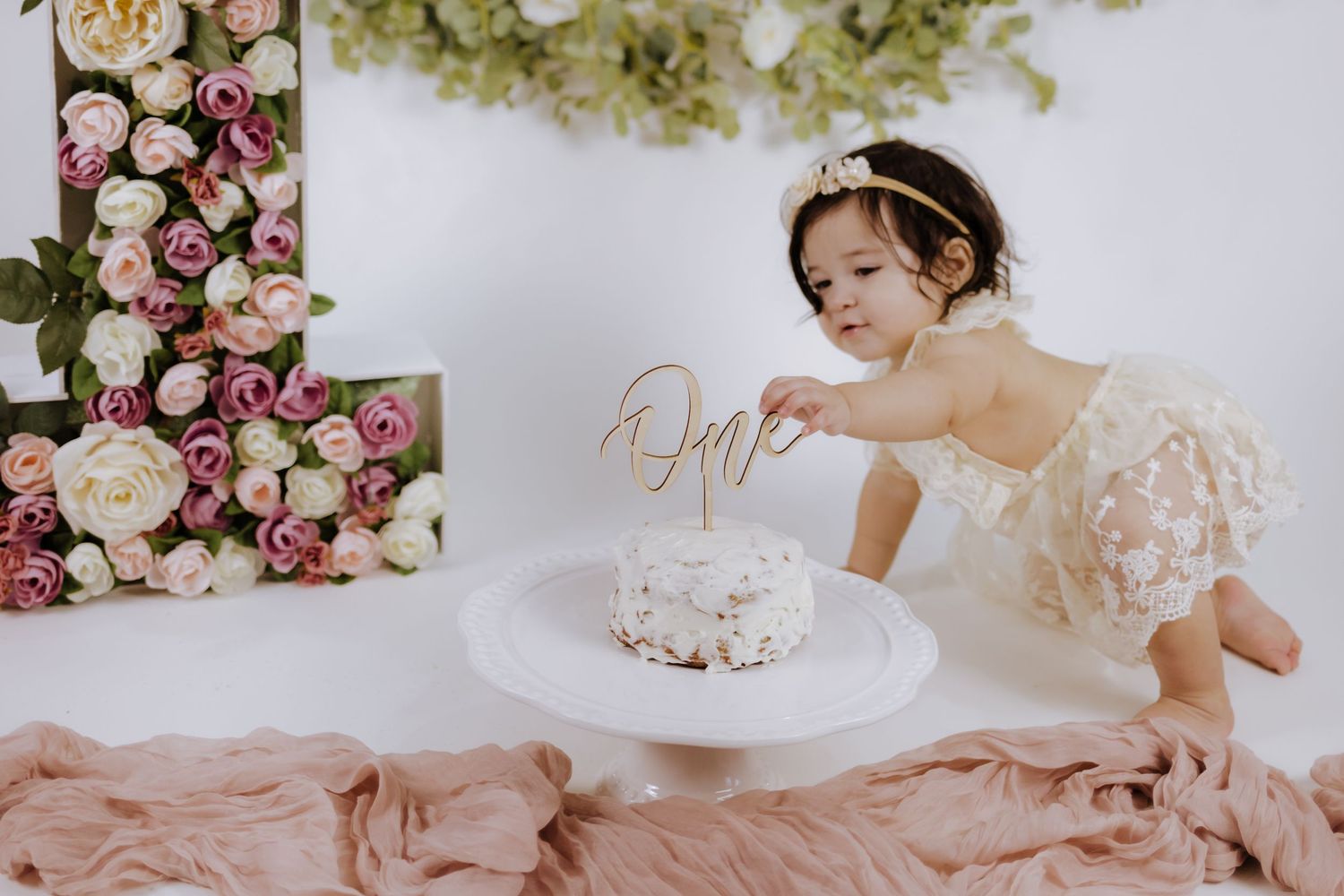 Baby girl in lace dress reaches for first birthday cake with floral backdrop and rose gold cake topper.
