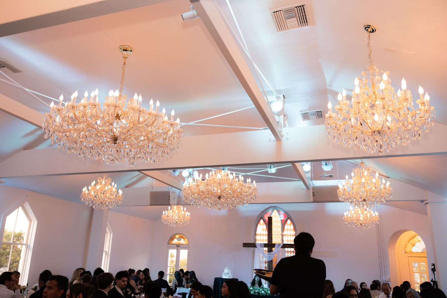 Elegant wedding reception hall with crystal chandeliers and arched windows filled with seated guests.