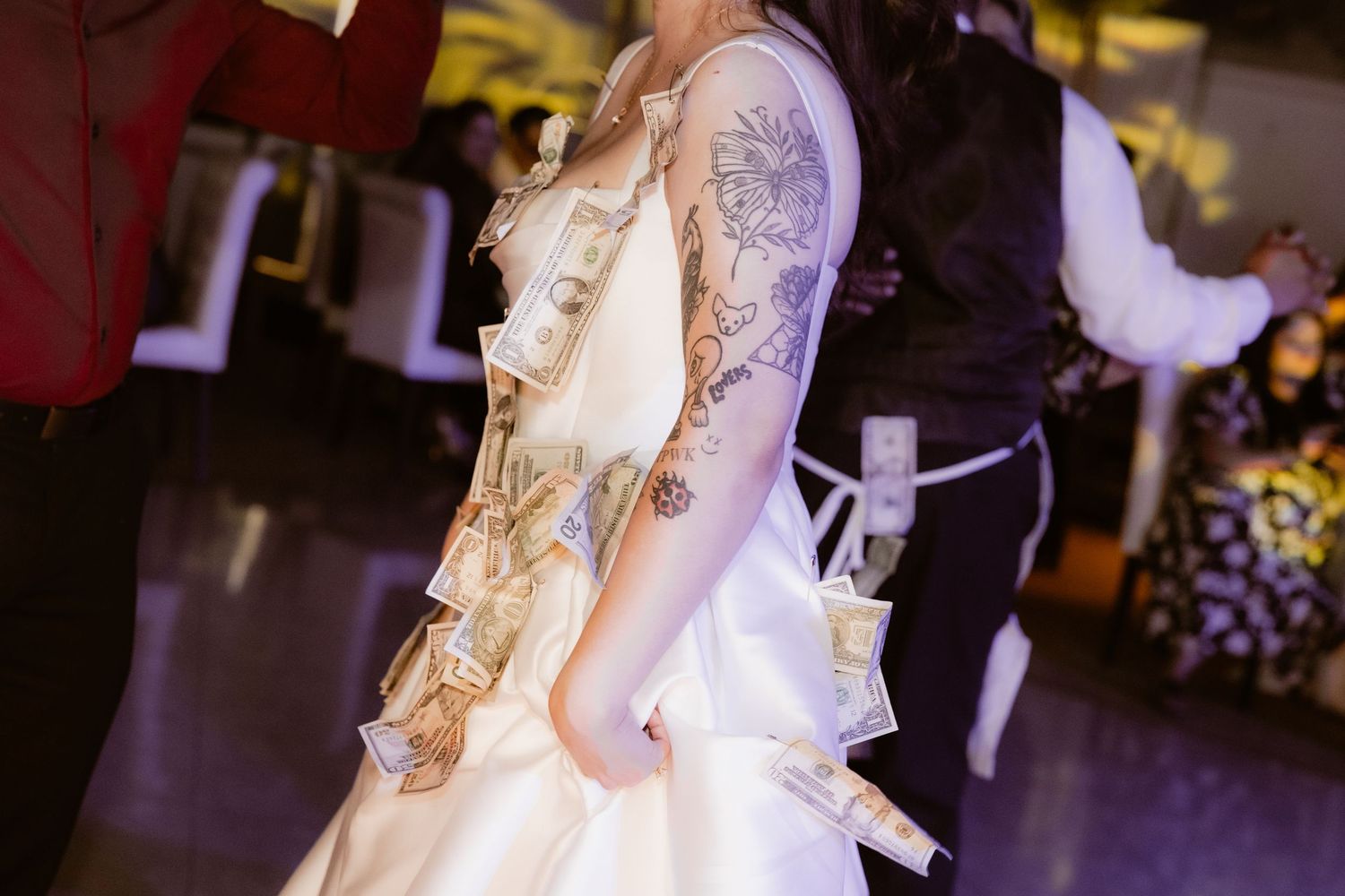 Bride in white satin gown with floral tattoo sleeve covered in dollar bills during reception dance.