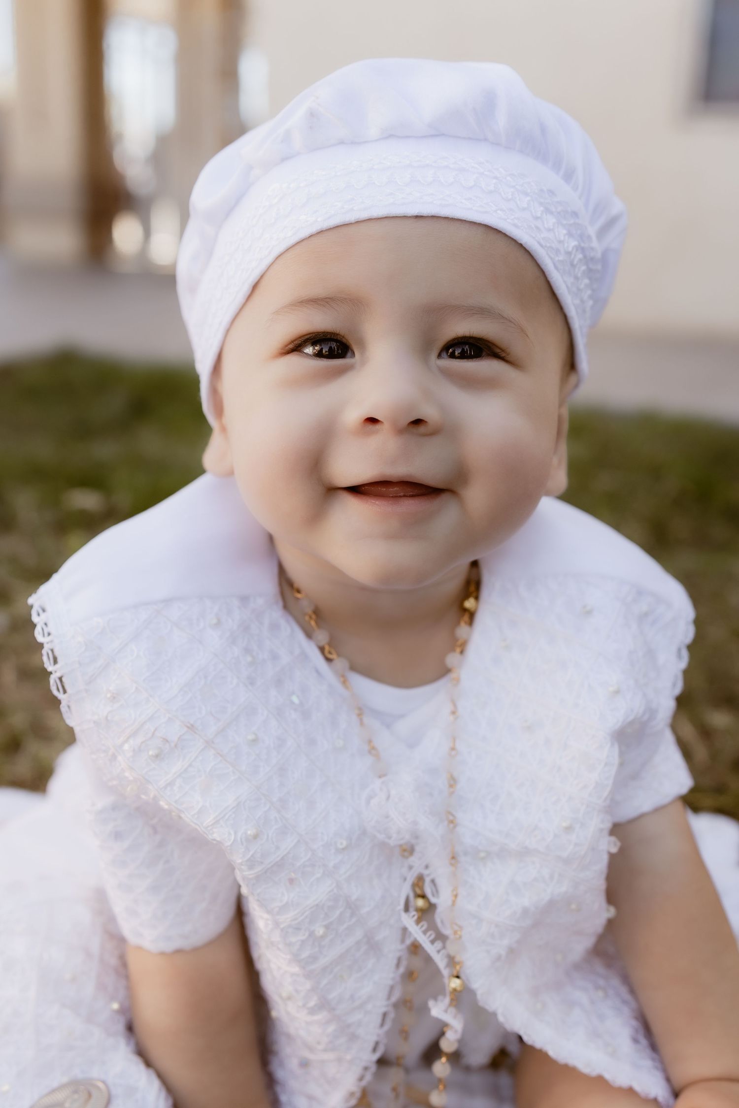 Smiling baby in white outfit and white hat wearing gold necklace outdoors.