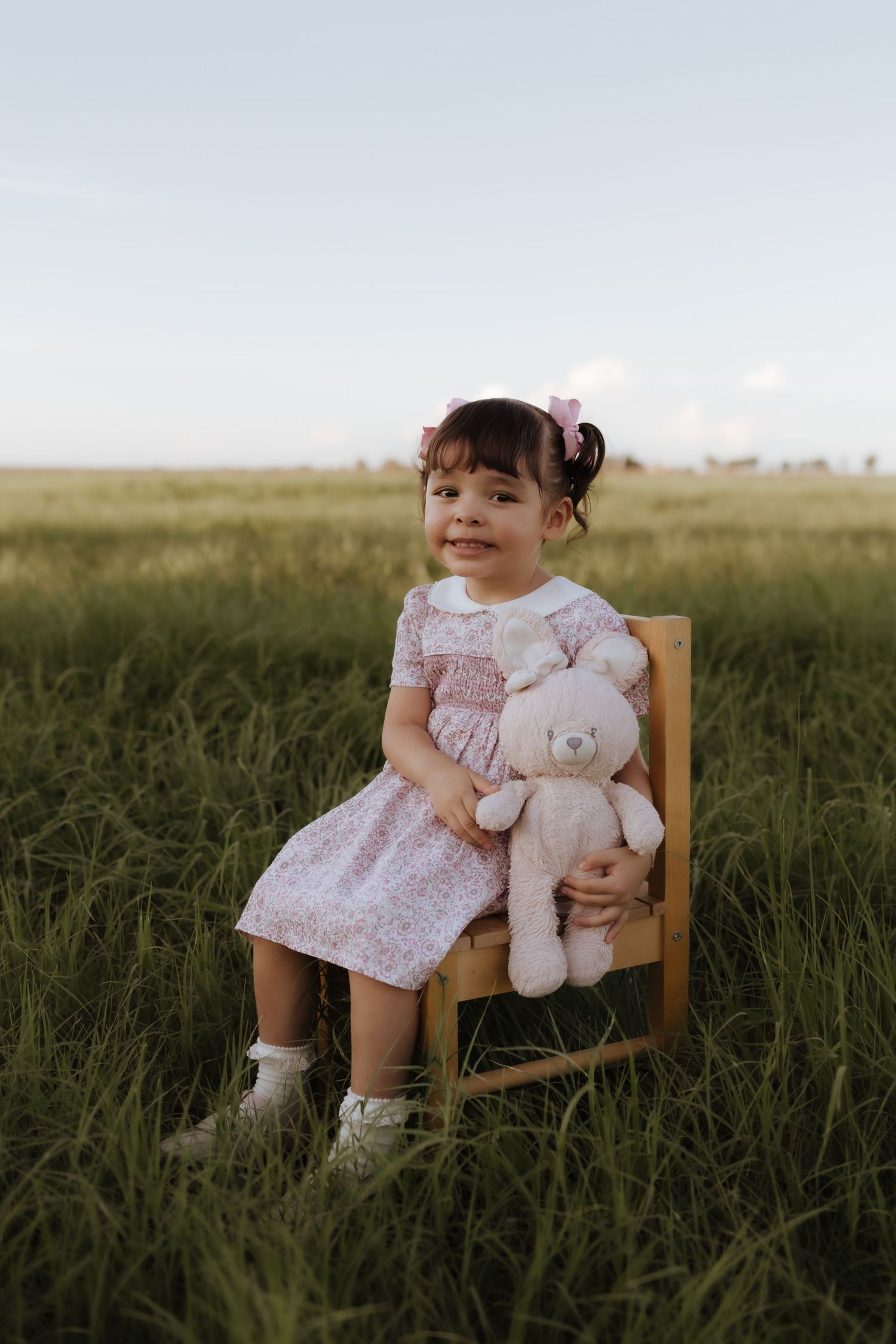 Toddler girl in floral pink dress sitting on wooden chair holding stuffed bear in green field.