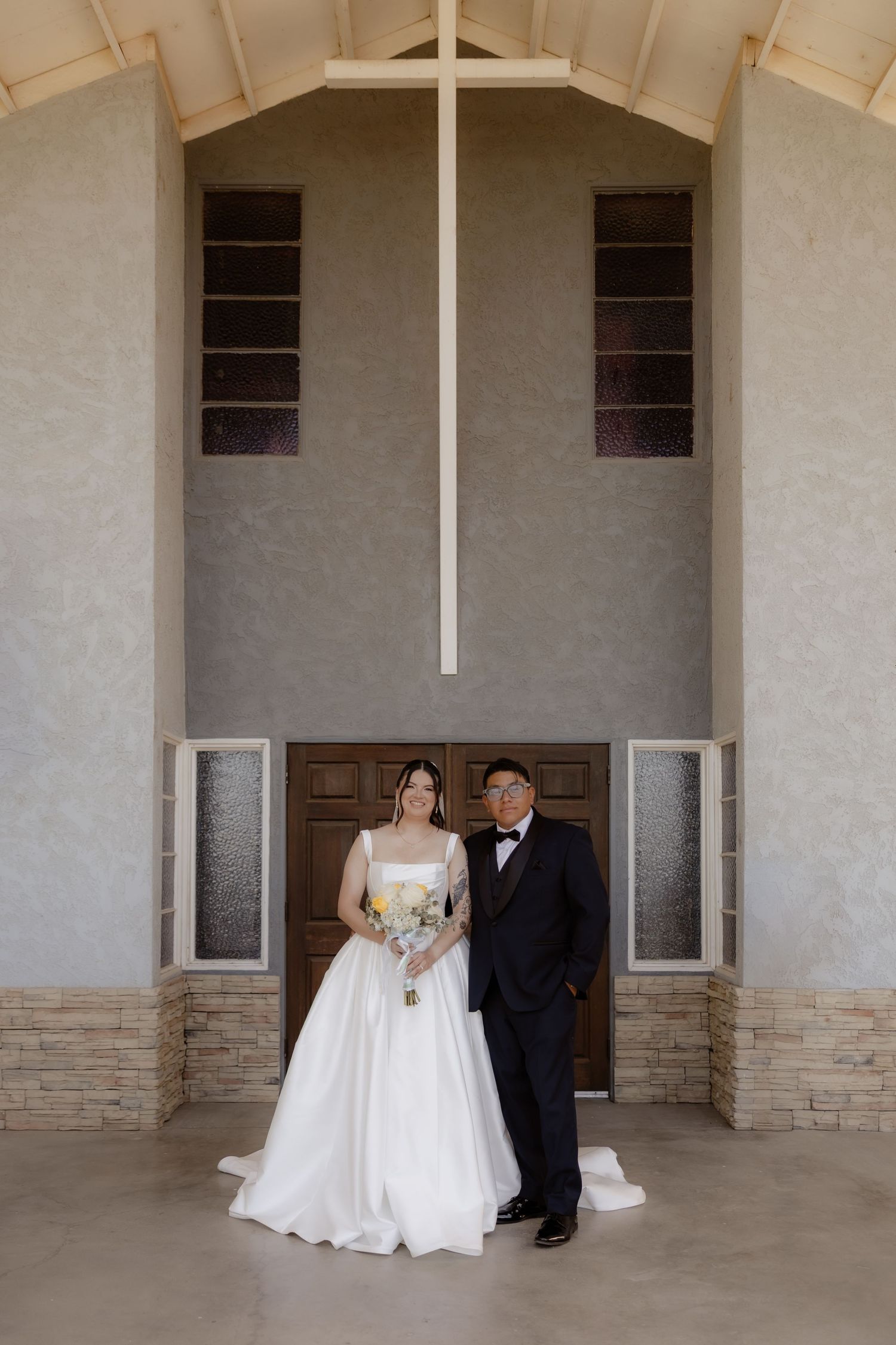Bride and groom stand together in front of large wooden cross inside a modern church chapel.