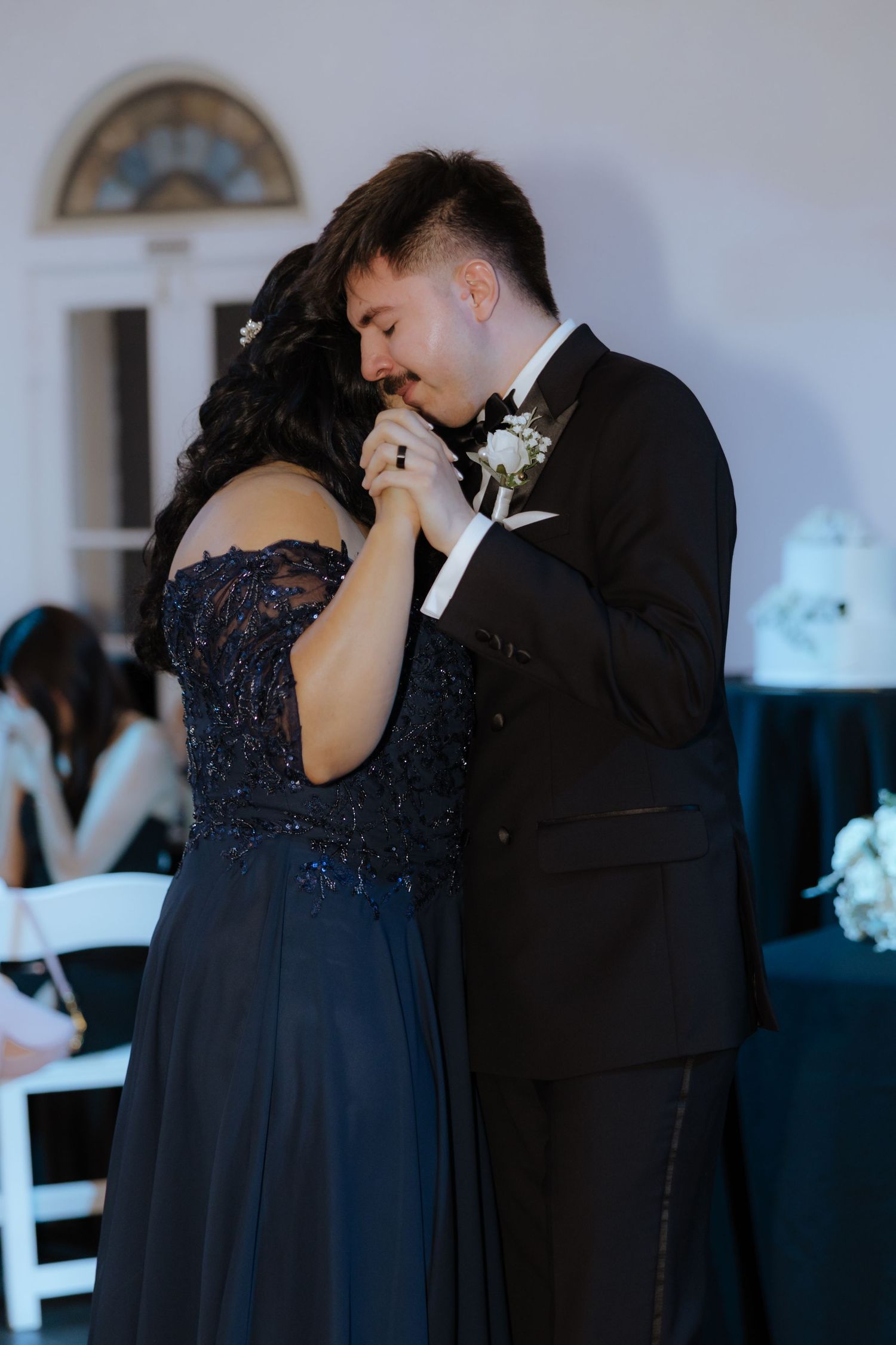 Groom in black tuxedo kissing bride's hand during an intimate mother-son dance at wedding reception.