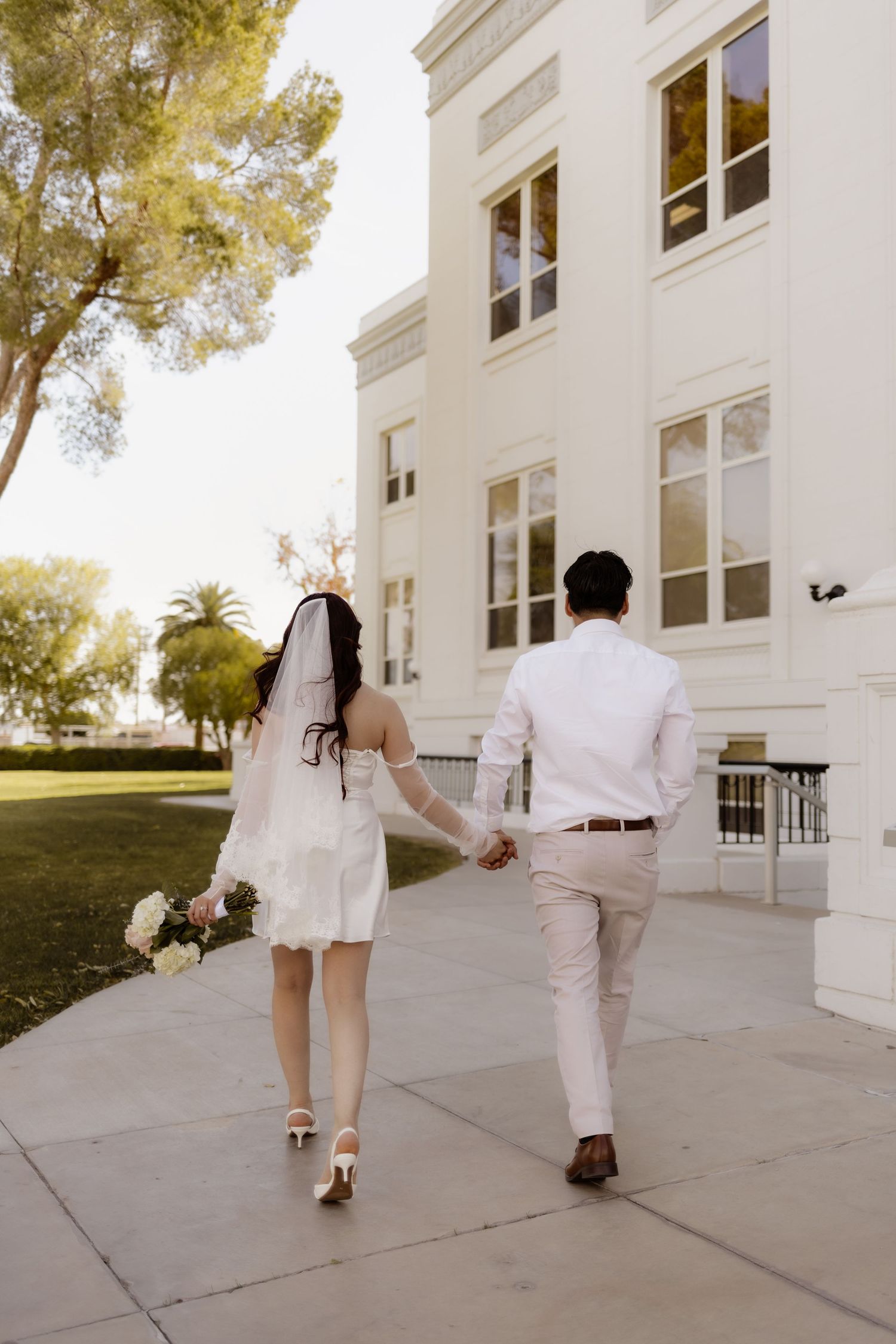 Bride and groom holding hands walking toward a white building on their wedding day.
