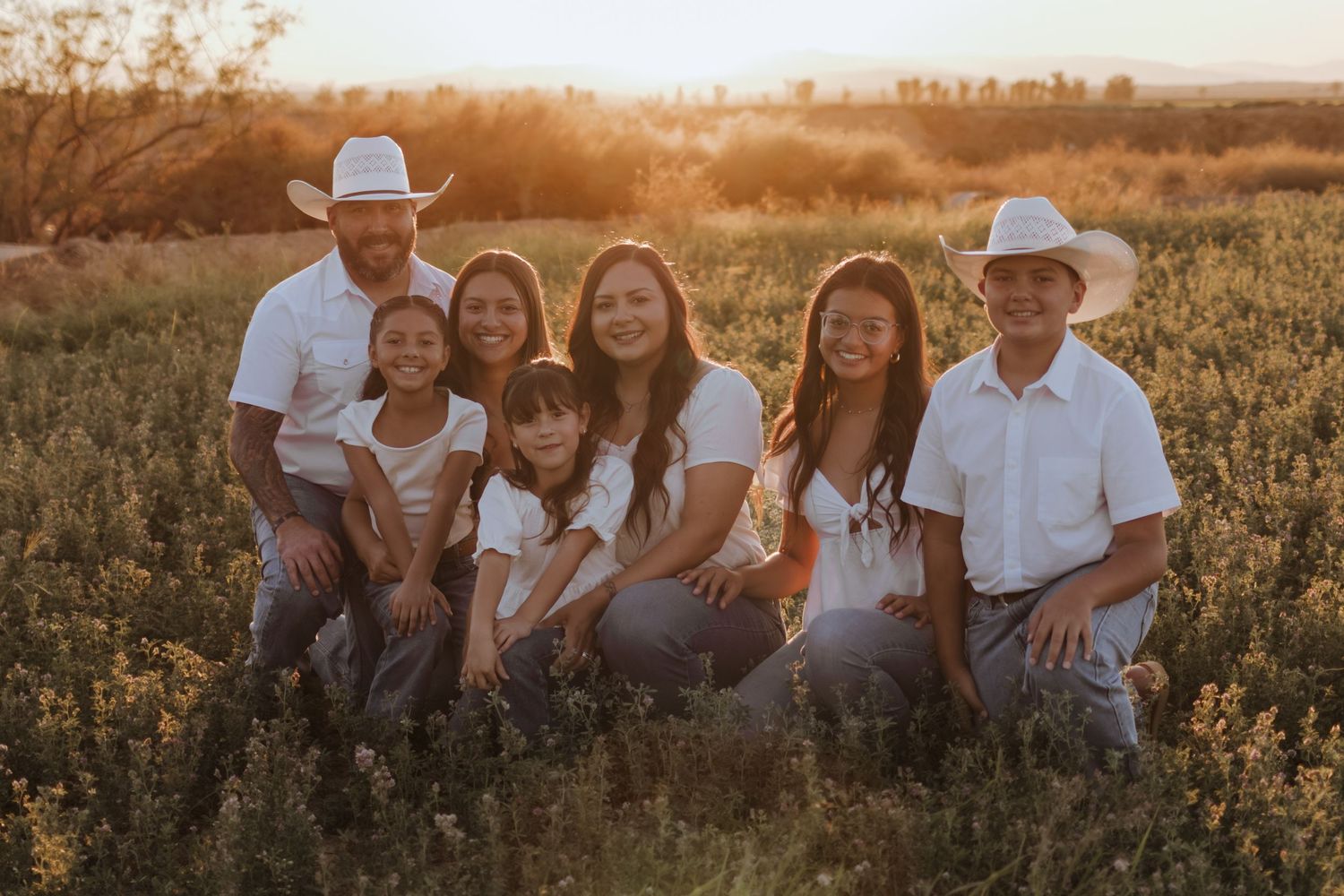 A family of seven in white shirts and cowboy hats poses together in a golden sunlit field.