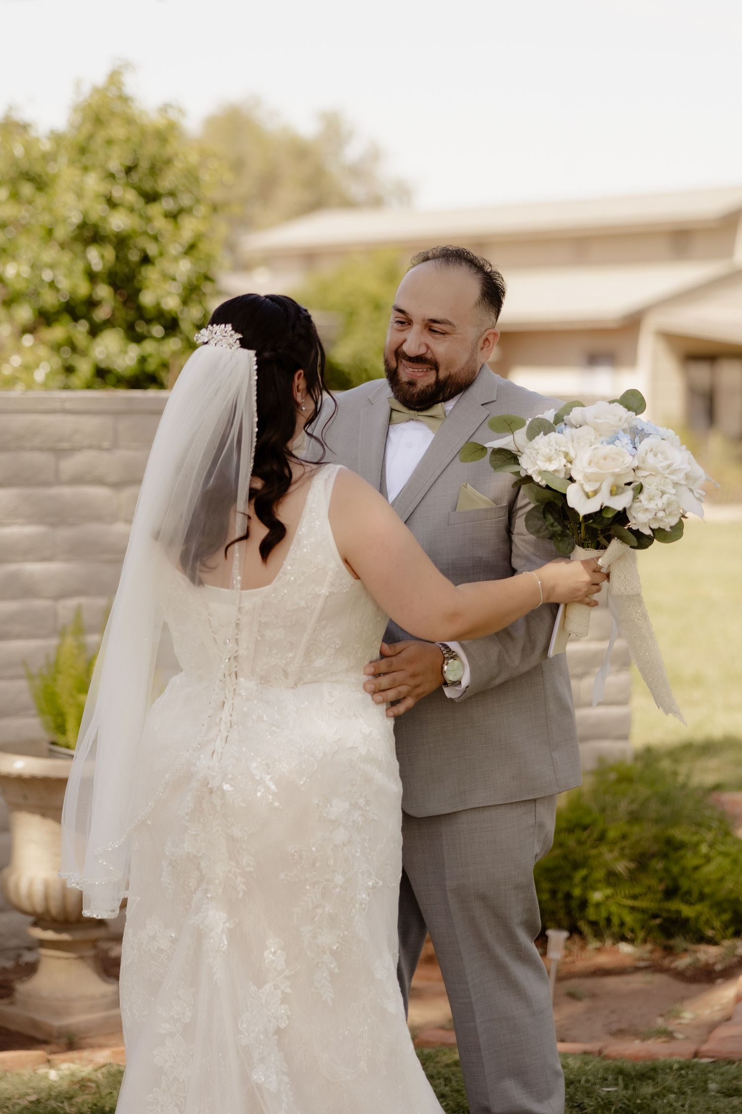 Groom in gray suit smiles joyfully at his bride during an outdoor first look moment while she holds white bouquet.