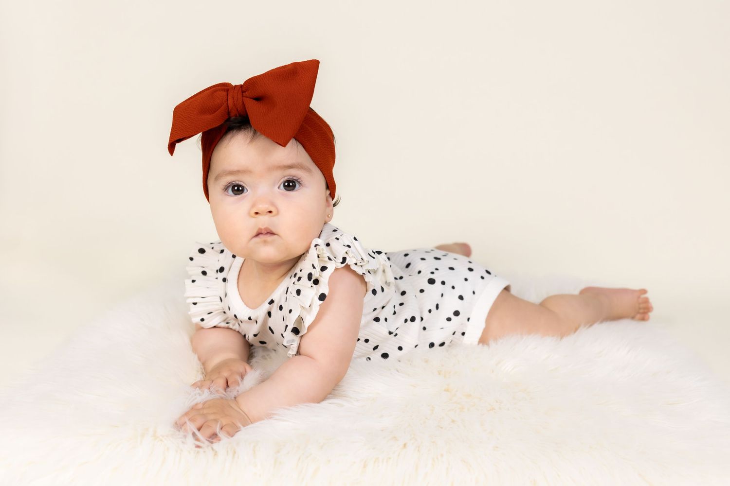 Adorable baby girl in polka dot romper and rust bow headband lying on white fur rug.