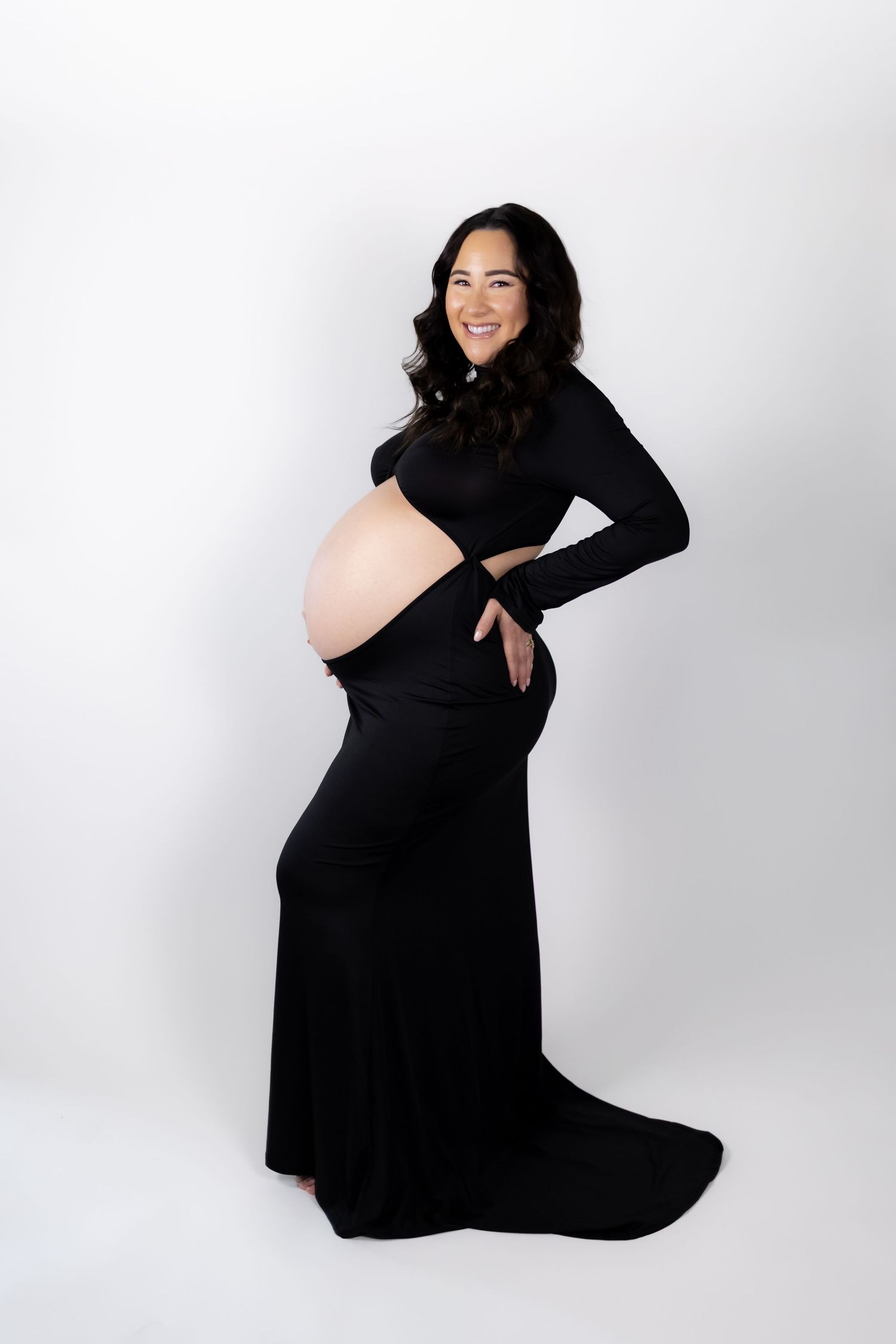 Smiling pregnant woman in black cutout maternity gown posing against white background in professional photo shoot.