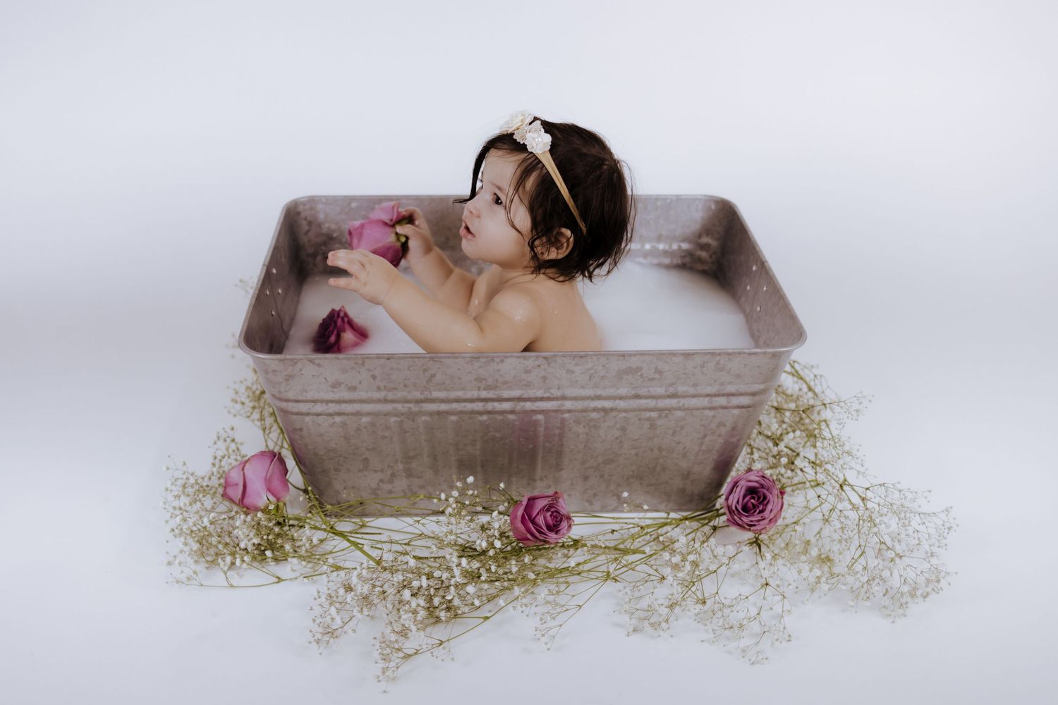Young toddler girl sitting in vintage metal tub with milk bath, pink roses and baby's breath flowers surrounding her.