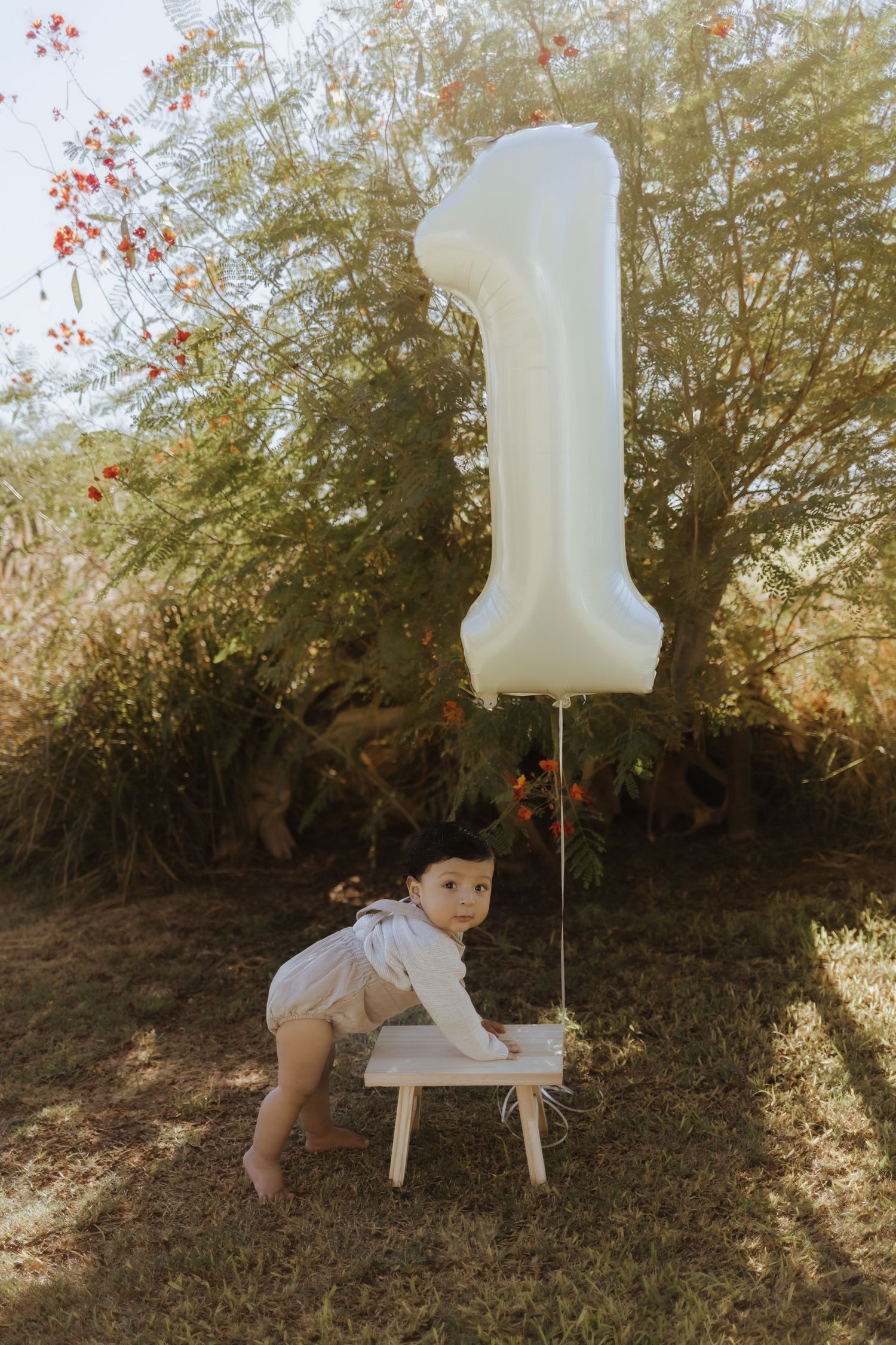 Baby in white outfit standing by number 1 balloon tied to small white stool outdoors.