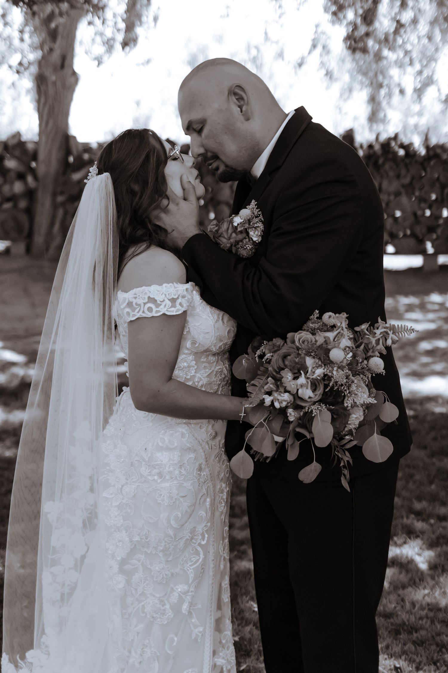 Black and white romantic portrait of bride and groom nearly kissing outdoors, groom holding a beautiful floral bouquet.