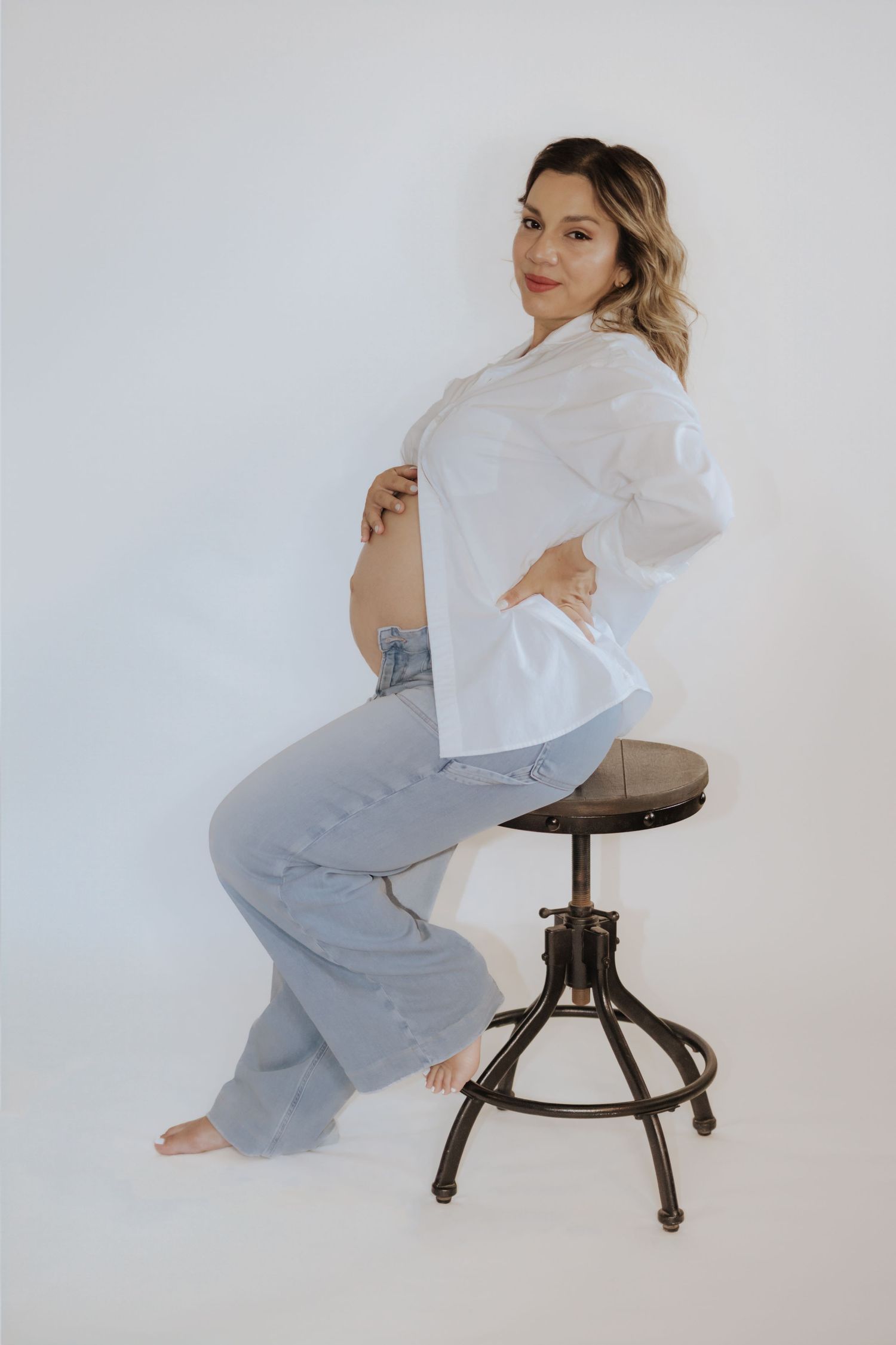 Pregnant woman in white shirt and light jeans sitting on industrial stool, cradling baby bump in studio setting.