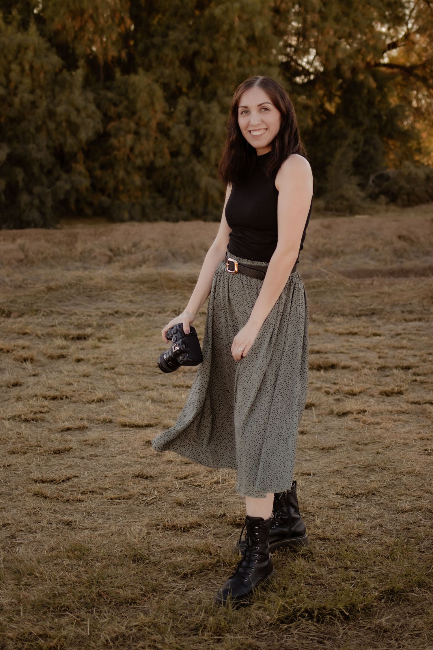 Female photographer smiling outdoors holding a camera, wearing black top and midi skirt.