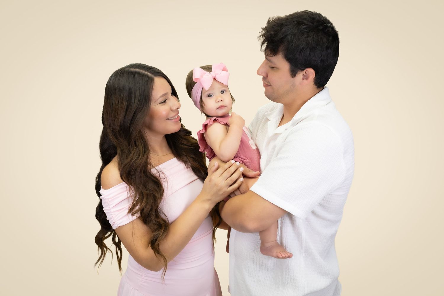 A young couple in pink and white outfits lovingly hold their baby girl in a pink dress and bow headband.