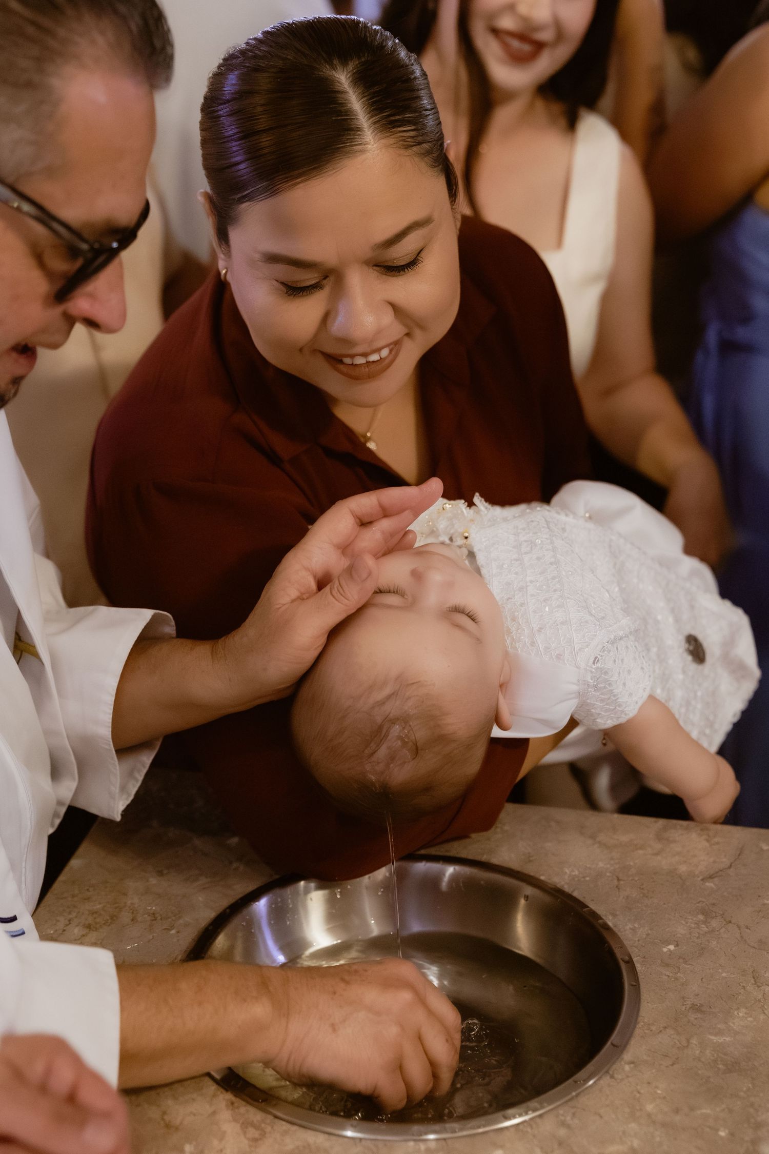 Priest baptizing infant over bowl while smiling woman in burgundy dress holds the baby.