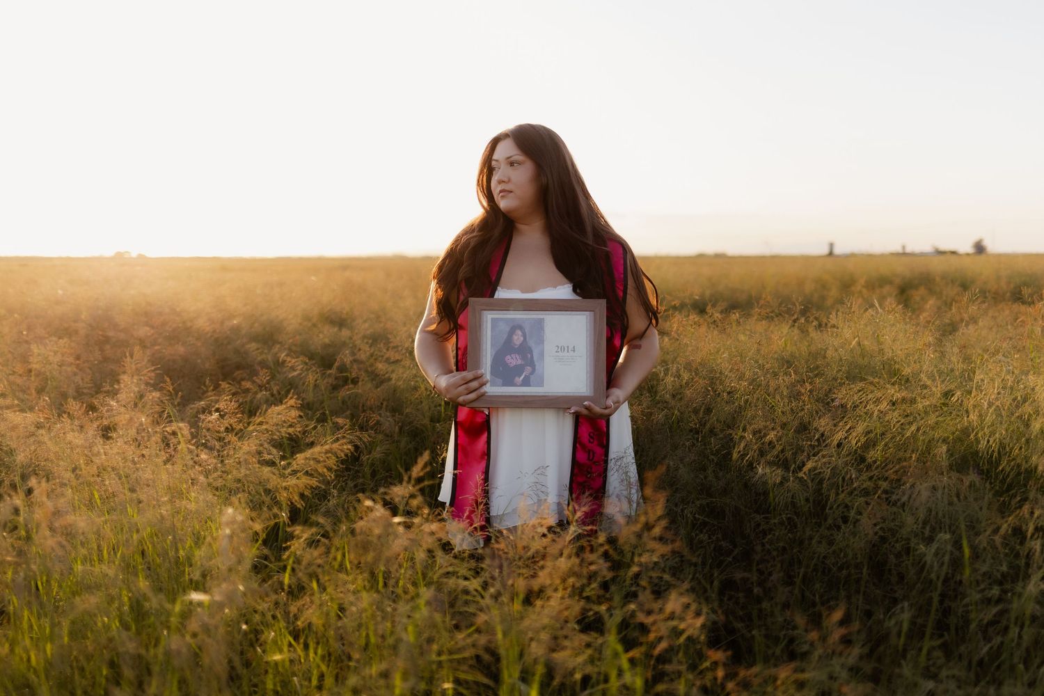 Young woman with long red hair holds framed photo while standing in golden field at sunset.