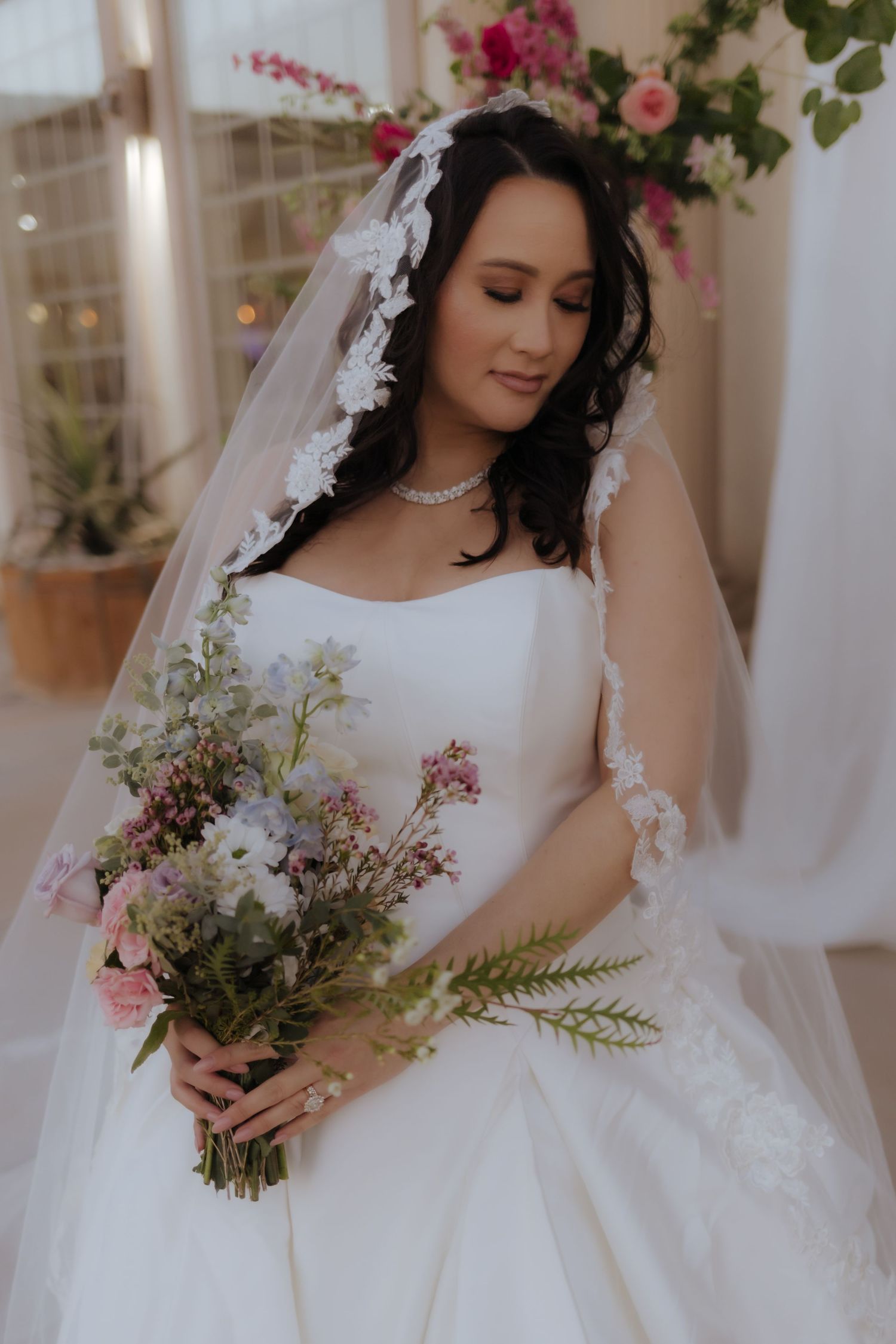 Brunette bride in white gown with lace veil holds pink and white floral bouquet in soft indoor light.