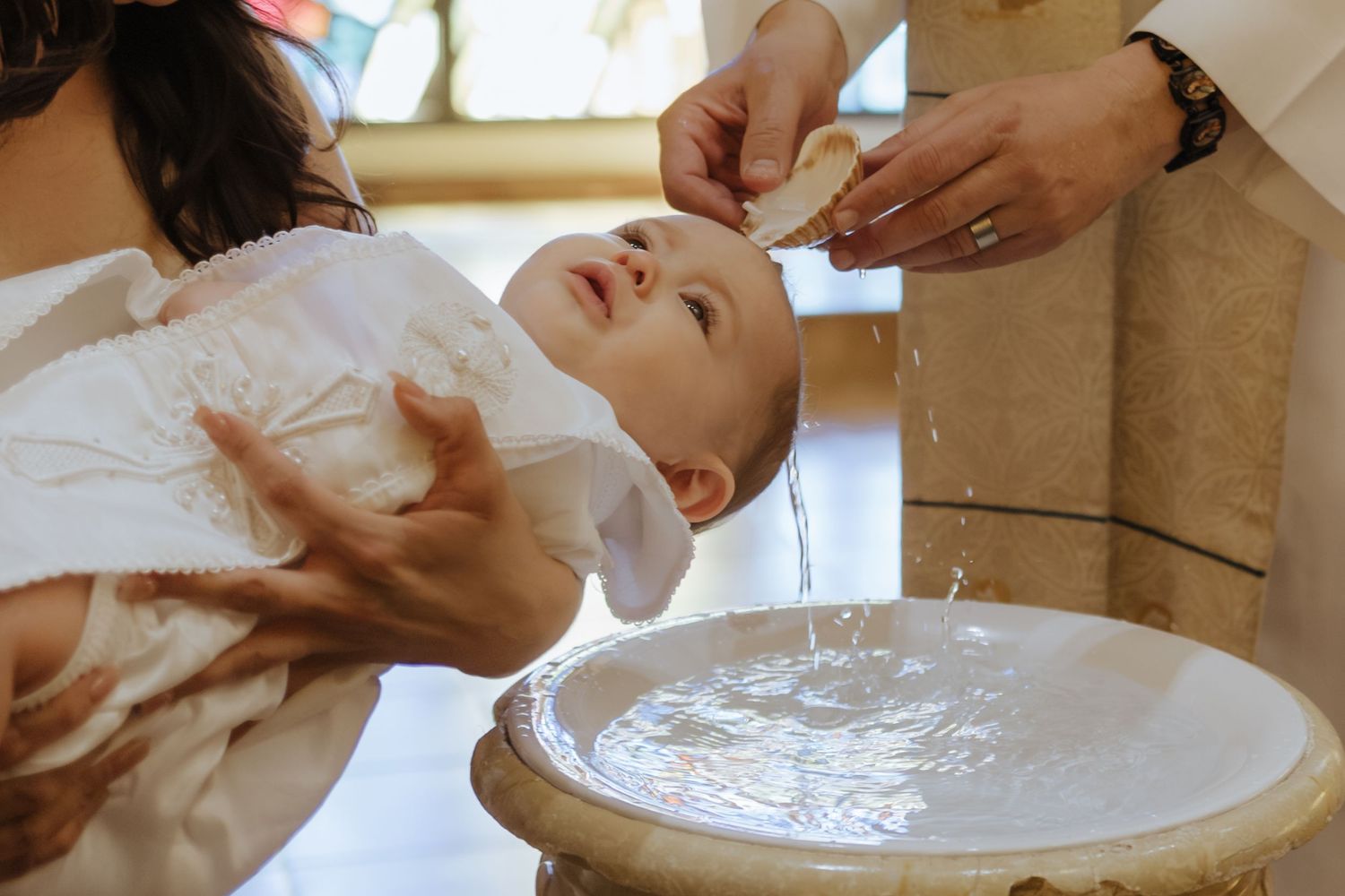 Baby being baptized with water poured over head at stone baptismal font in church.