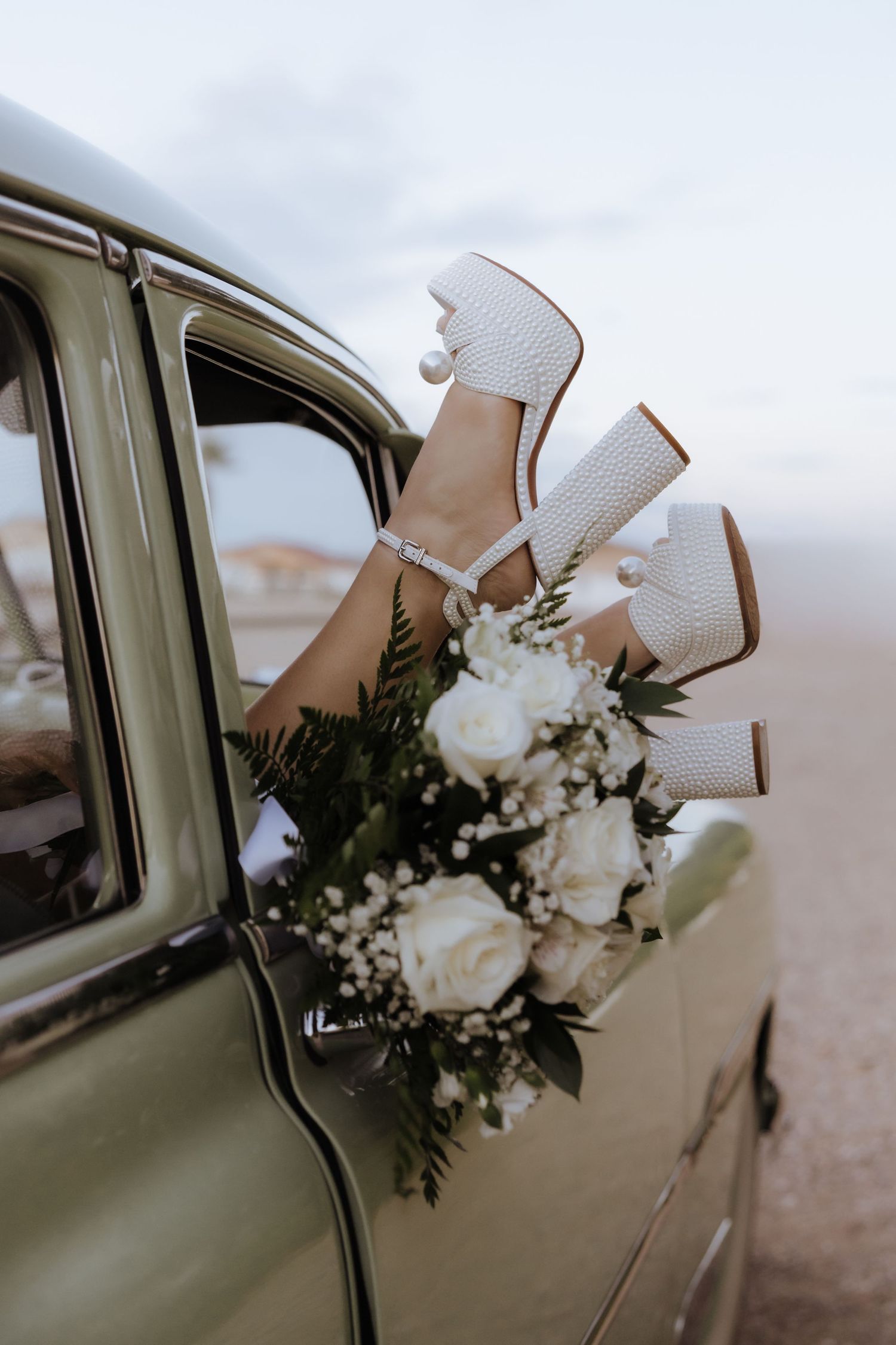 Brides white platform heels and wedding bouquet of white roses extending from a vintage car window.