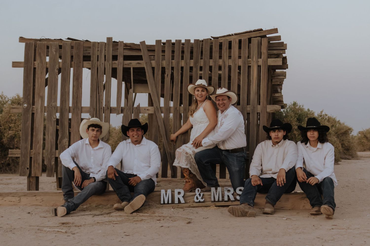 A couple and their group pose around a Mr. and Mrs. sign at a rustic outdoor desert setting at dusk.