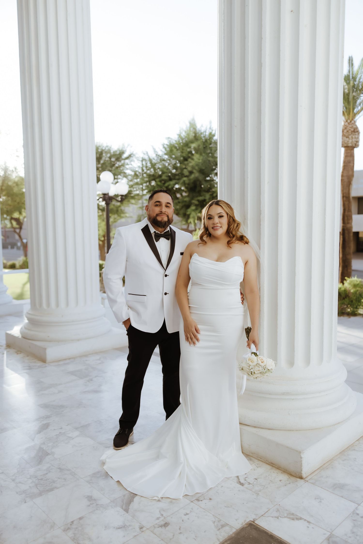 Bride and groom posing between white columns in elegant wedding attire with bouquet.
