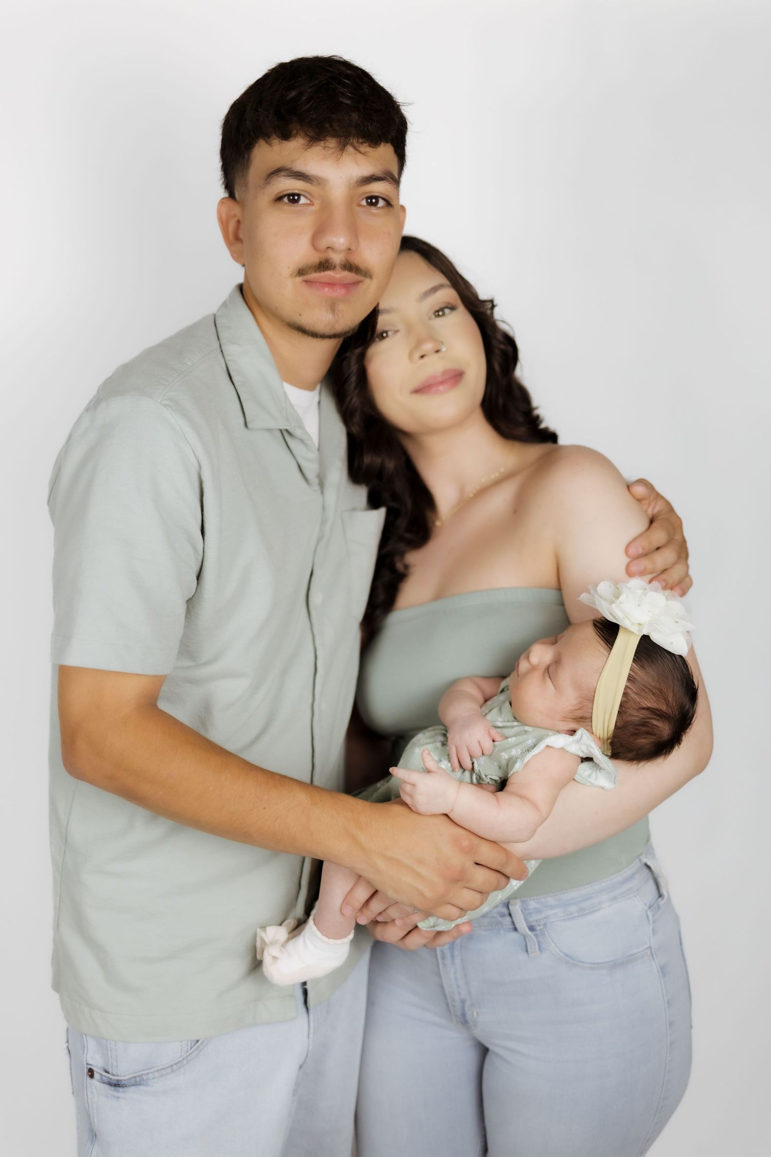 Young couple in matching sage green outfits tenderly holding their newborn baby against a white background.