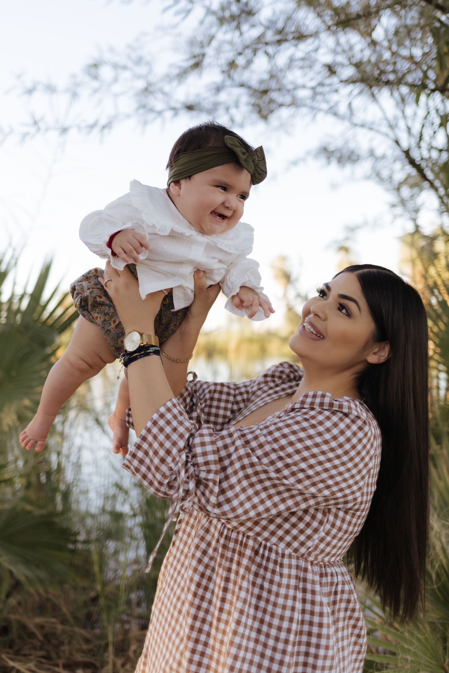 Smiling mother lifts happy baby girl in white dress outdoors near palm trees.