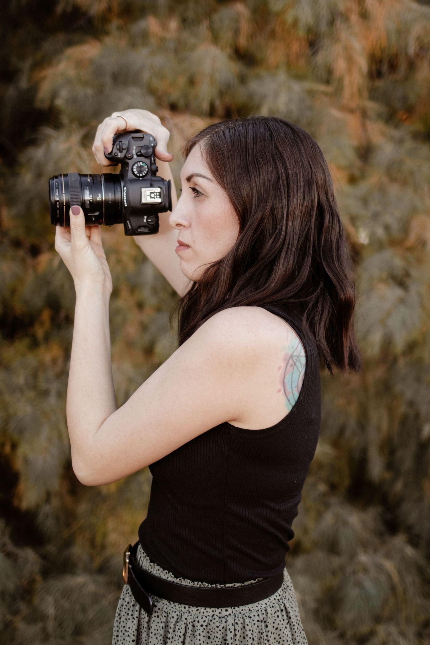 Female photographer with tattoo raising DSLR camera to shoot, standing near pine trees.