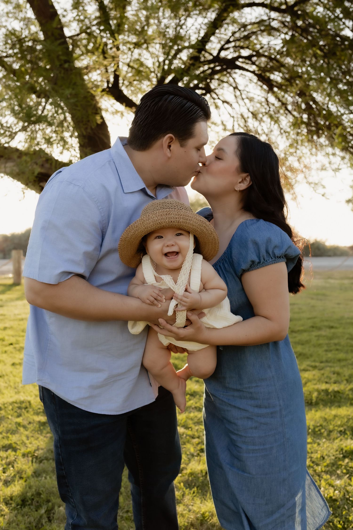 Happy couple kissing while holding laughing baby in straw hat outdoors in golden sunlight.