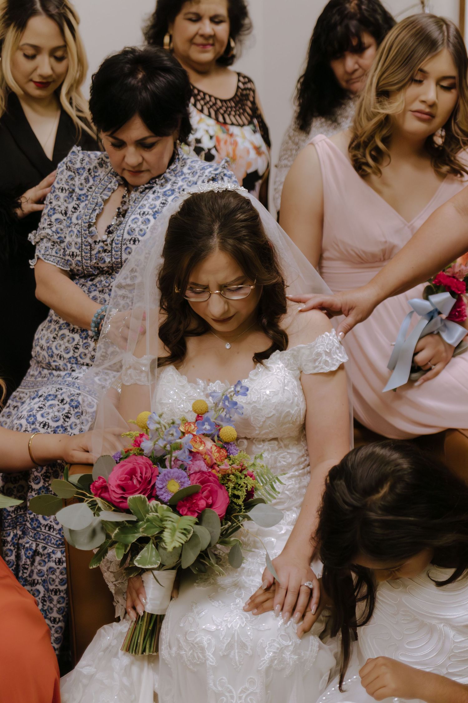 Bride wearing glasses holds colorful bouquet of pink and purple flowers surrounded by bridesmaids in blush pink dresses.