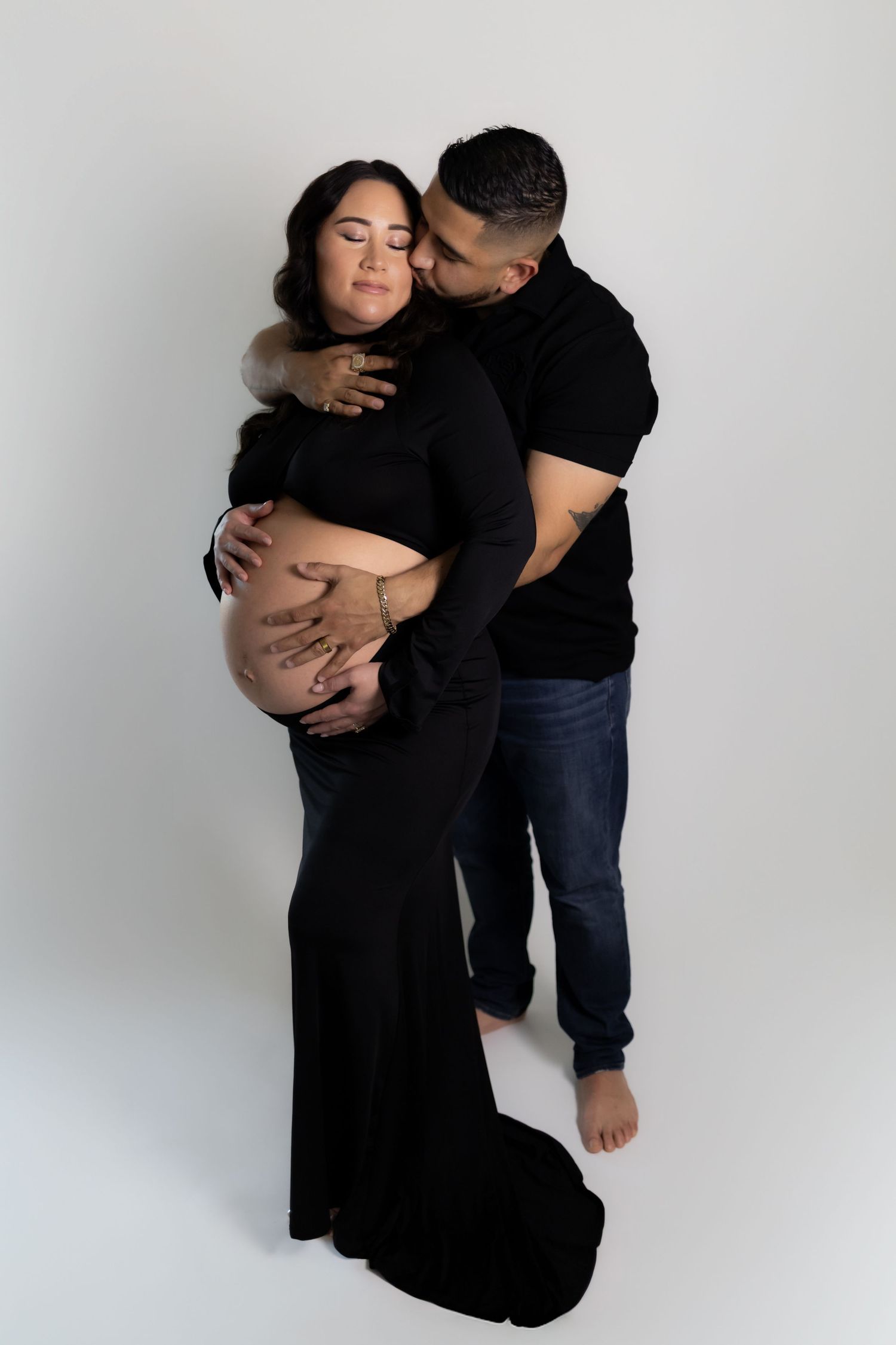 Couple in black outfits posing for maternity photo, man embracing pregnant woman from behind on white background.