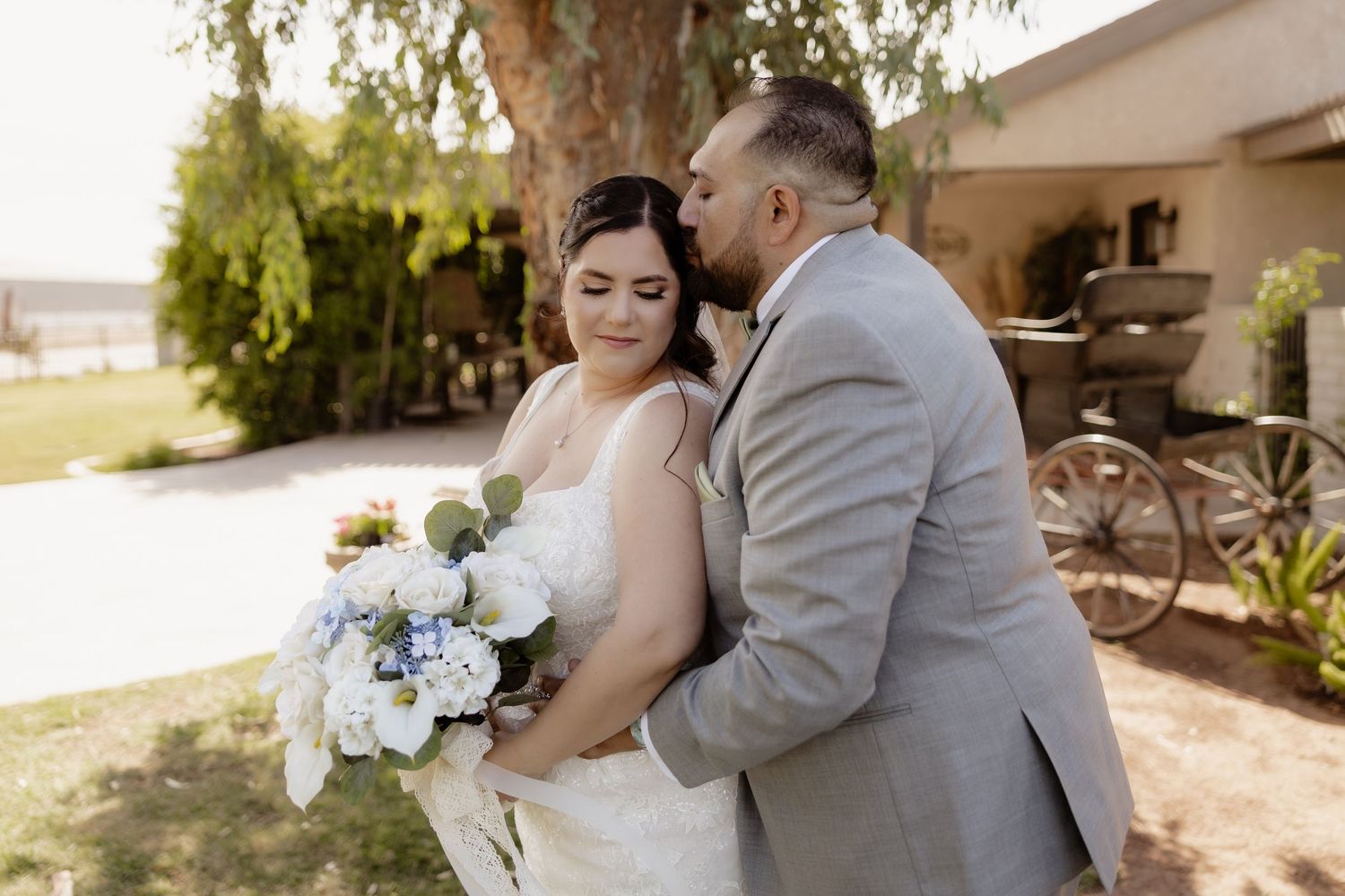 Groom in gray suit kissing bride on cheek as she holds white and blue floral bouquet outdoors.