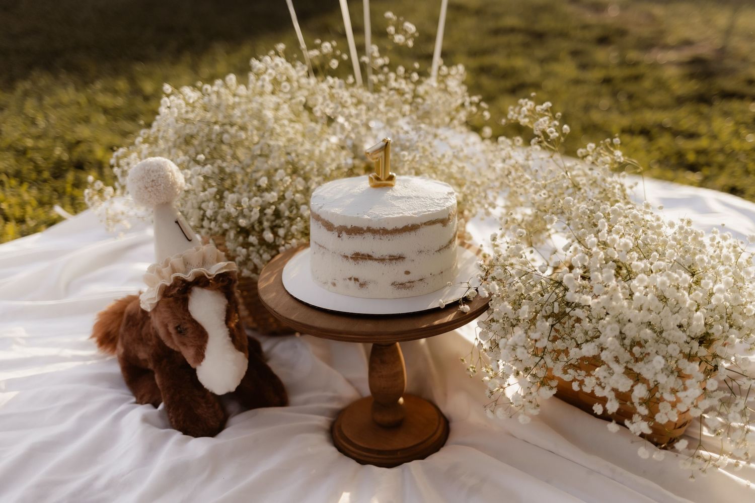 First birthday smash cake on wooden stand surrounded by baby's breath flowers and stuffed animal.