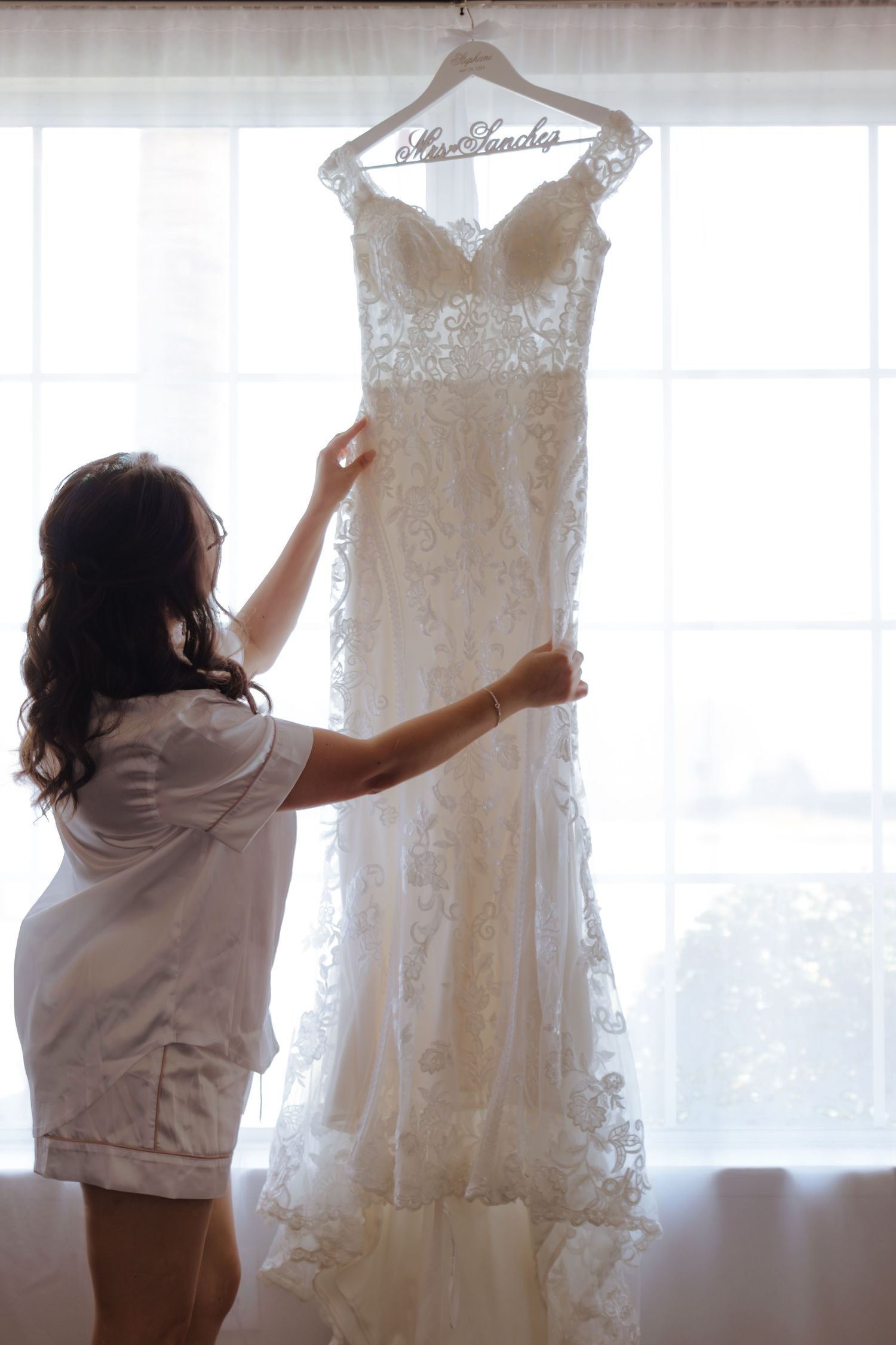 Bride in white robe admires her lace wedding gown hanging on a personalized Mrs. Sanchez hanger by a bright window.