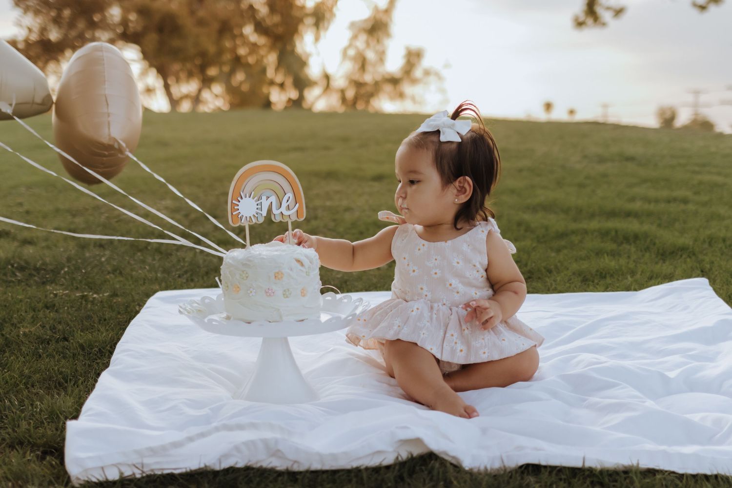 Baby girl in floral dress reaching for first birthday cake with rainbow topper outdoors.