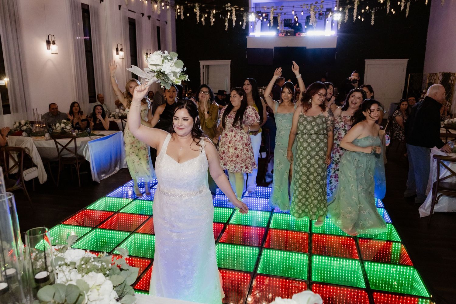 Bride raises white bouquet triumphantly on a colorful LED dance floor surrounded by cheering guests at wedding reception.