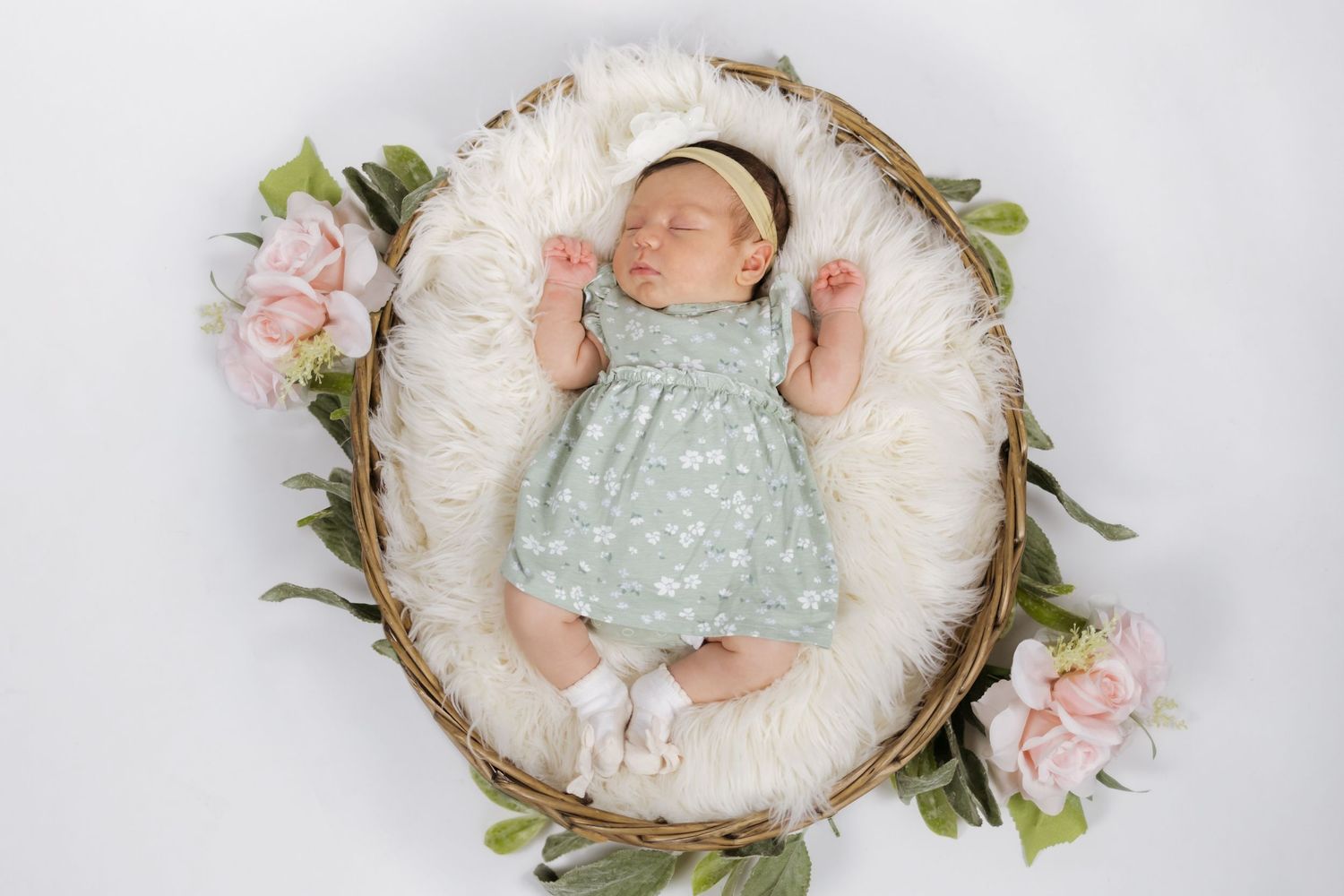 Newborn baby girl sleeping in a wicker basket with white fur and pink flowers wearing a mint green dress.