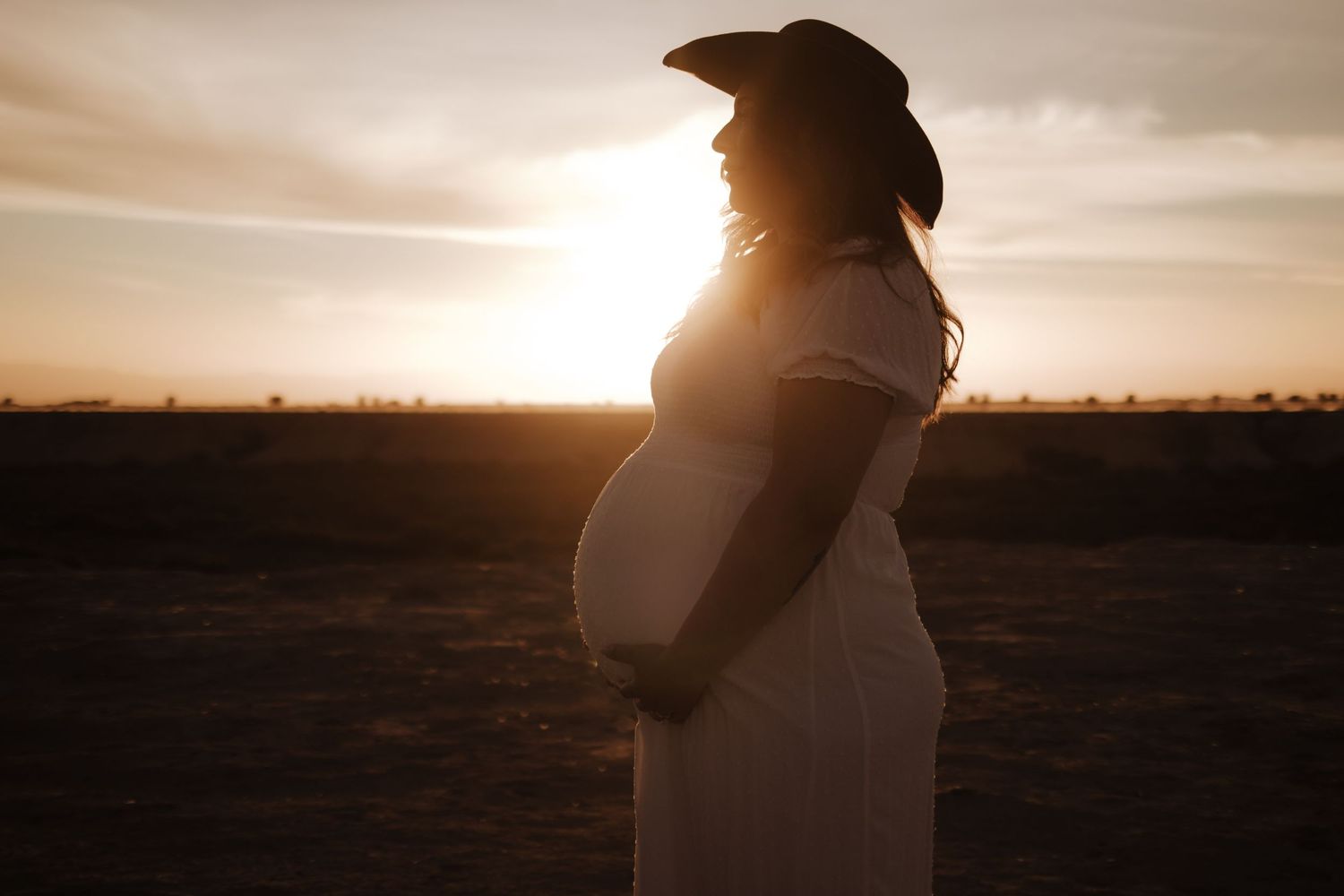 Silhouette of pregnant woman in white dress and cowboy hat at golden sunset outdoors.