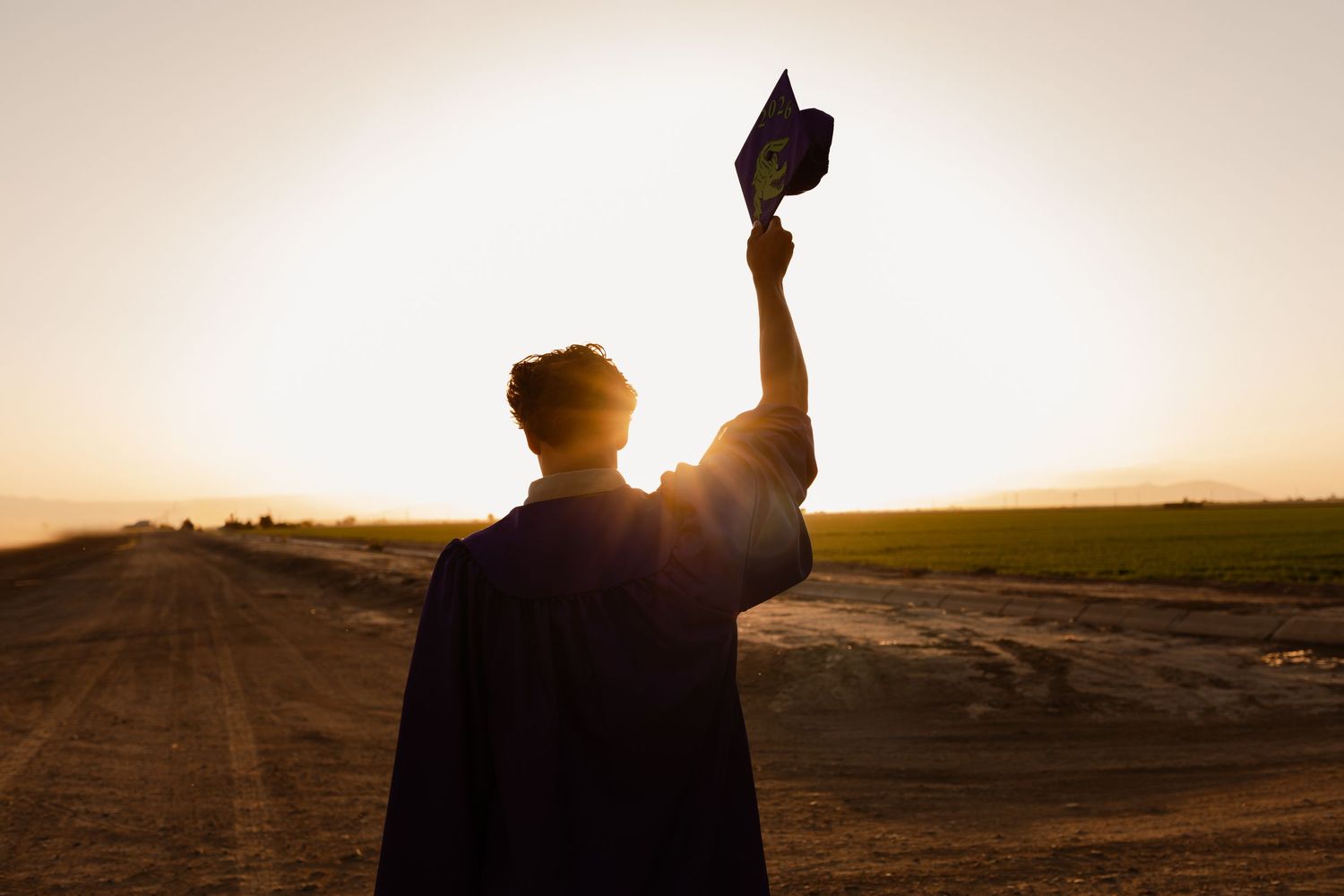 Graduate in cap and gown silhouetted against golden sunset raising mortarboard in the air.