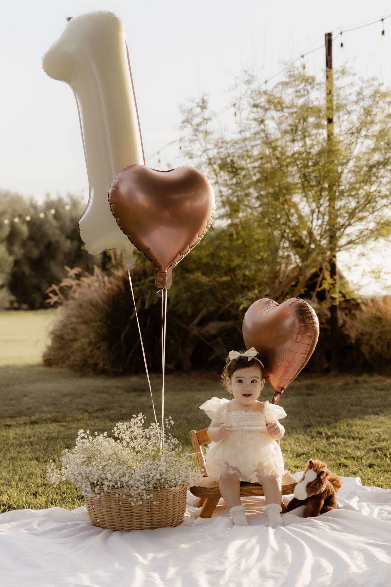Baby girl in white dress sitting with heart balloons and number 1 balloon outdoors.
