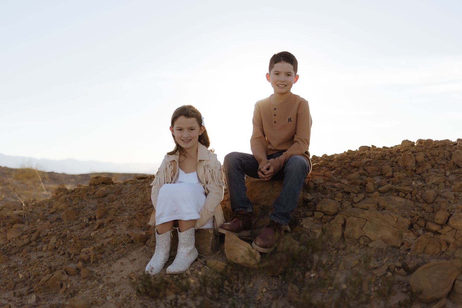 A young girl and boy sit together on rocky terrain with a clear sky and desert landscape behind them.