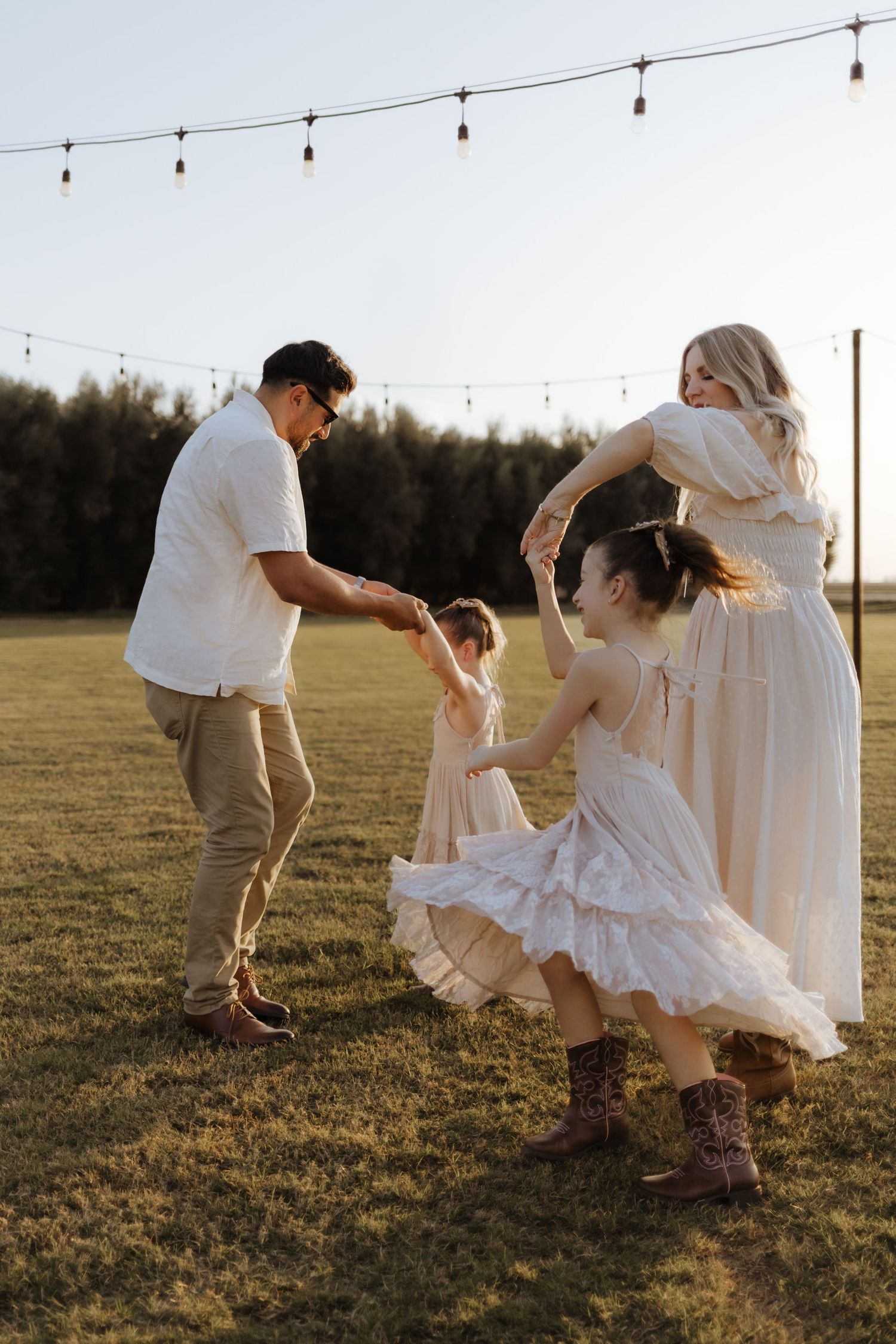 Family dancing together in a sunlit field with string lights overhead at golden hour.