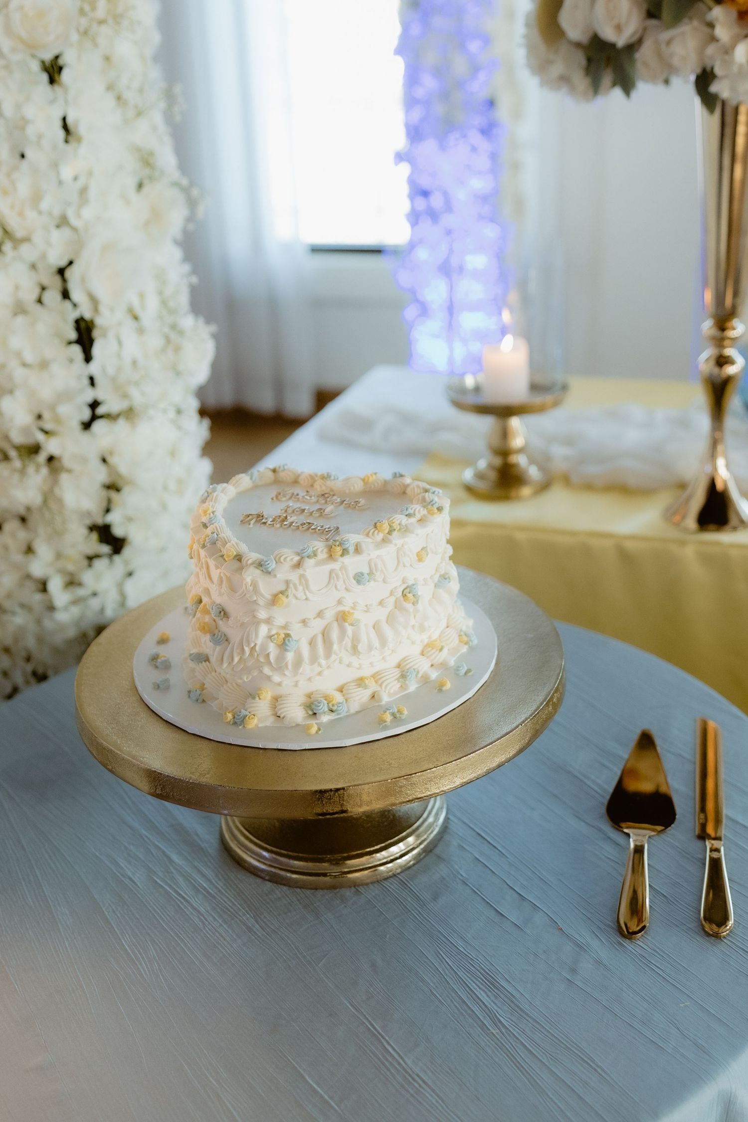 Elegant single-tier white wedding cake with colorful sprinkles on a gold cake stand surrounded by white floral arrangements.