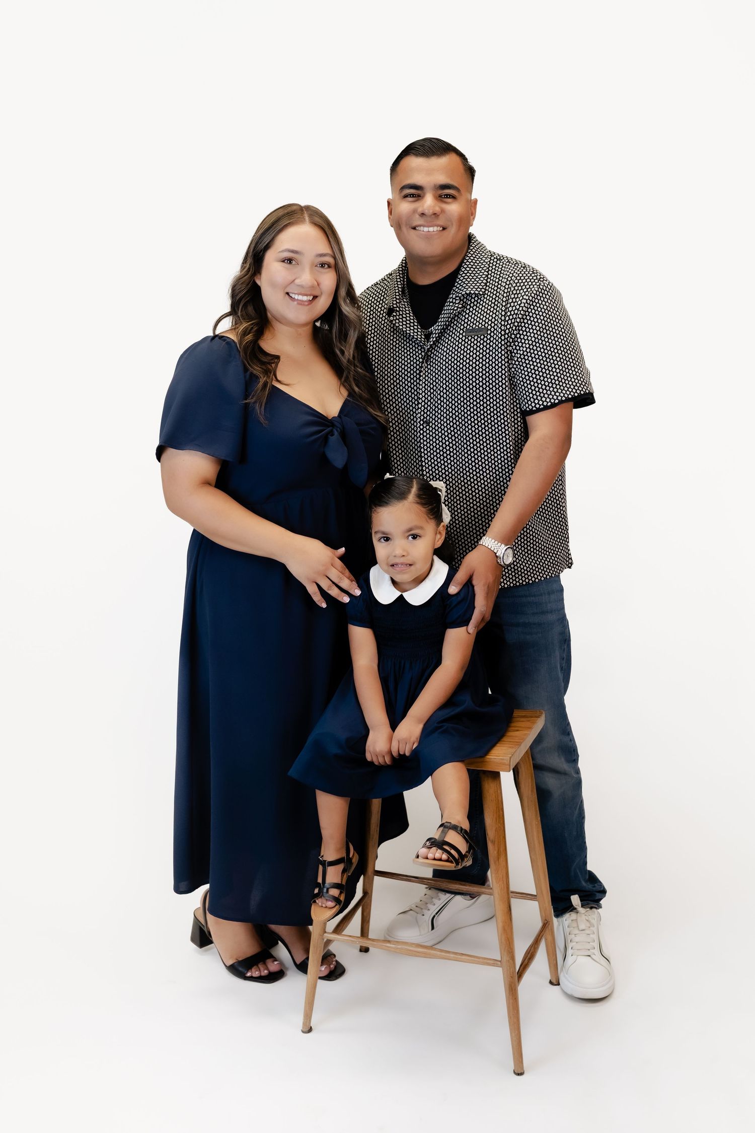 A smiling family of three poses in navy blue outfits against a white background, with a toddler girl seated on a stool.