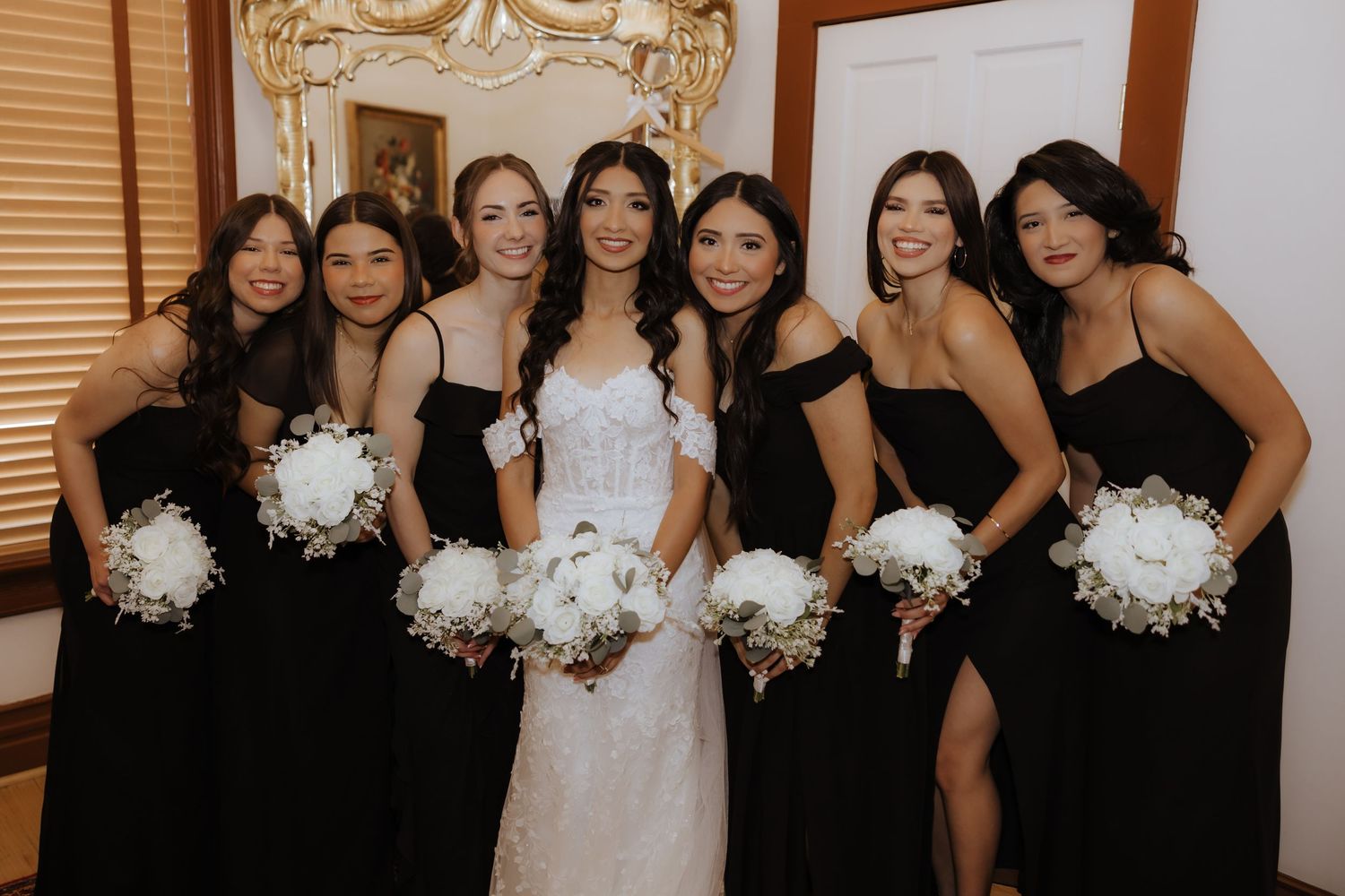 Bride in white gown posing with six bridesmaids in black dresses holding white peony bouquets indoors.