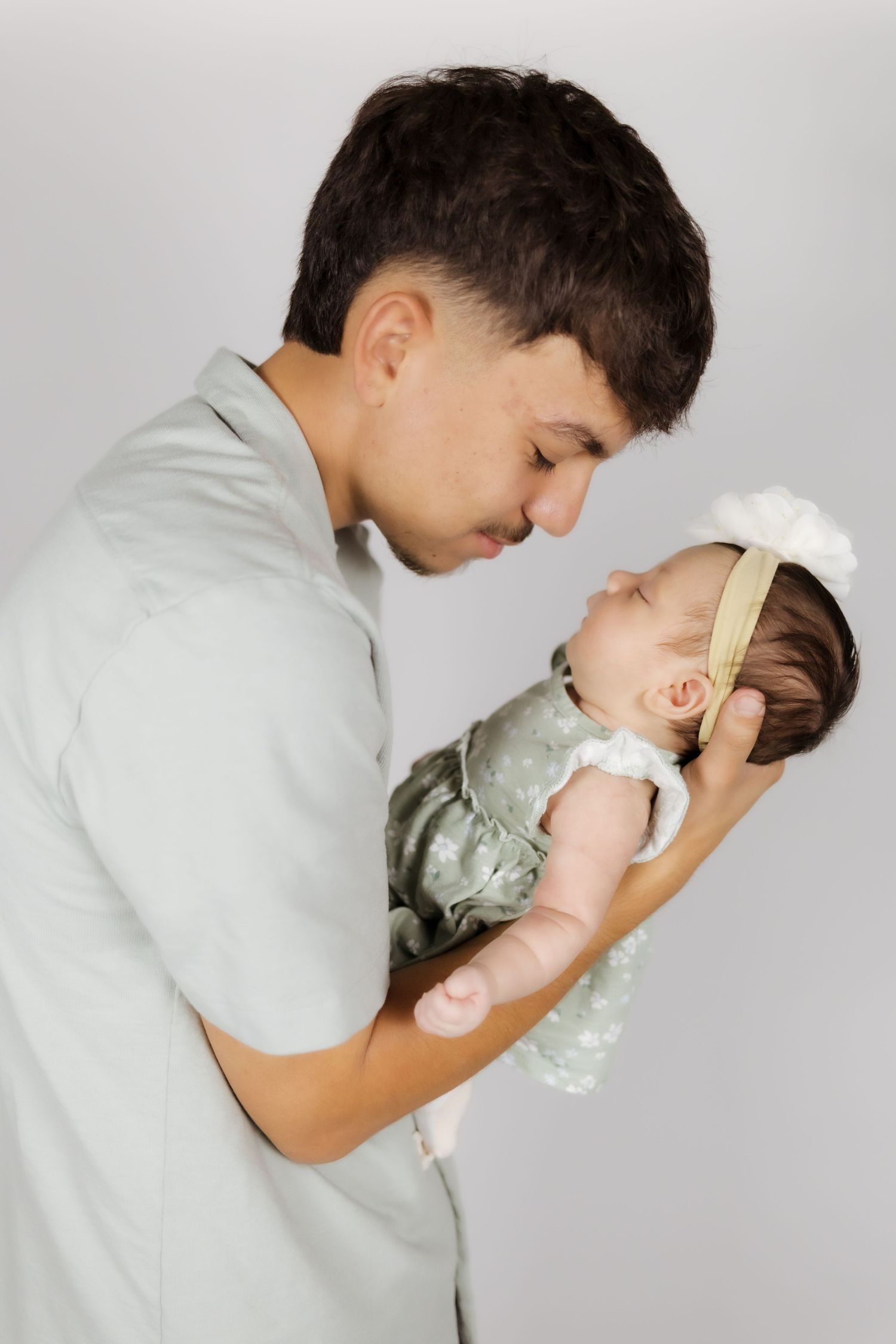 Young father tenderly holding sleeping newborn baby girl in white bow headband against gray background.
