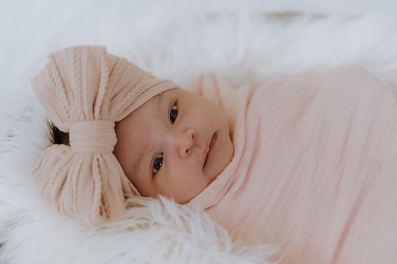 Newborn baby girl swaddled in pink wrap wearing a bow headband, lying on white fluffy blanket.